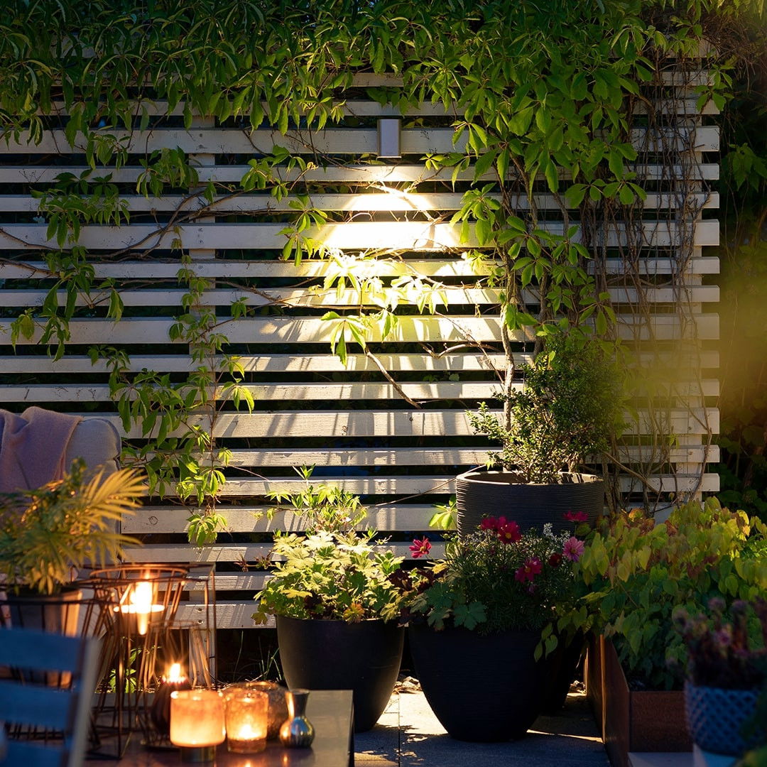 This image showcases a beautifully designed outdoor living space that seamlessly integrates natural and artificial elements to create a warm and inviting atmosphere. The focal point is the wooden lattice structure, which is partially obscured by the lush, verdant foliage that cascades over the slats. The strategic placement of the lighting fixtures within the lattice casts a warm, ambient glow, illuminating the surrounding plants and creating a cozy, intimate ambiance. The potted plants and flowers in the foreground add pops of vibrant color and texture, further enhancing the natural aesthetic. The combination of the wooden lattice, the greenery, and the soft lighting creates a visually striking and harmonious composition. The overall design suggests a thoughtful and intentional approach to blending the built and natural elements, resulting in a captivating and inviting outdoor retreat. The attention to detail, such as the positioning of the lighting and the selection of the plant materials, contribute to the overall sense of refinement and sophistication within this carefully curated space. This image exemplifies the transformative power of thoughtful landscape and lighting design, where the boundaries between the indoor and outdoor environments are seamlessly dissolved, allowing the user to fully immerse themselves in a visually stunning and serene setting.