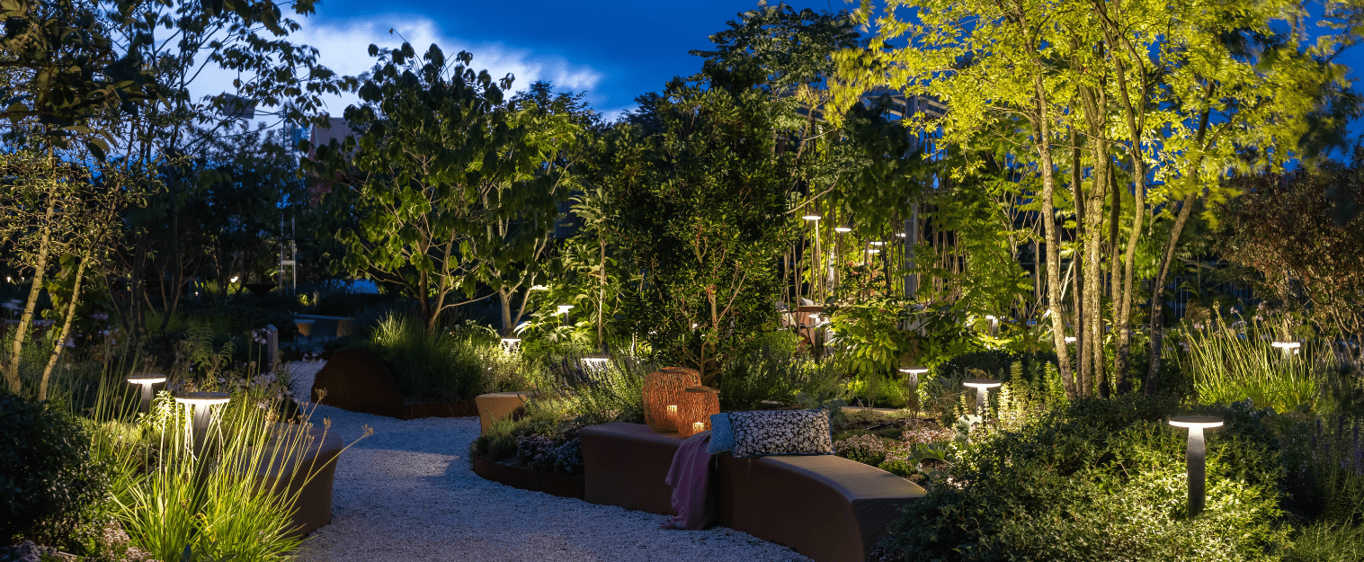 This image depicts a beautifully designed and illuminated outdoor garden scene at night. The focal point is a winding, stone pathway that meanders through a lush, verdant landscape filled with a variety of plants, trees, and landscape features. The pathway is lined with strategically placed lighting fixtures, creating a warm and inviting ambiance that highlights the vibrant colors and textures of the surrounding foliage. The lighting creates a serene and enchanting atmosphere, casting a soft glow over the entire scene. Surrounding the pathway are ornamental planters, benches, and other design elements that complement the natural setting. The use of natural materials, such as stone and wood, integrates the hardscaping seamlessly with the living plant life. In the distance, the silhouettes of tall trees and shrubs can be seen, creating depth and a sense of enclosure within the garden. The overall composition and attention to detail result in a visually stunning and immersive outdoor environment, inviting the viewer to explore and experience the tranquility and beauty of this carefully curated space. The image conveys a sense of harmony between the natural and the designed, with the thoughtful use of lighting and landscape elements creating a truly captivating and inviting outdoor oasis.