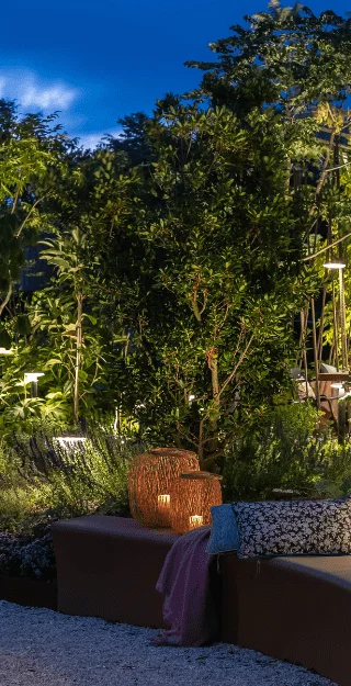 This image depicts a beautifully designed and illuminated outdoor garden scene at night. The focal point is a winding, stone pathway that meanders through a lush, verdant landscape filled with a variety of plants, trees, and landscape features.  The pathway is lined with strategically placed lighting fixtures, creating a warm and inviting ambiance that highlights the vibrant colors and textures of the surrounding foliage. The lighting creates a serene and enchanting atmosphere, casting a soft glow over the entire scene.  Surrounding the pathway are ornamental planters, benches, and other design elements that complement the natural setting. The use of natural materials, such as stone and wood, integrates the hardscaping seamlessly with the living plant life.  In the distance, the silhouettes of tall trees and shrubs can be seen, creating depth and a sense of enclosure within the garden. The overall composition and attention to detail result in a visually stunning and immersive outdoor environment, inviting the viewer to explore and experience the tranquility and beauty of this carefully curated space.  The image conveys a sense of harmony between the natural and the designed, with the thoughtful use of lighting and landscape elements creating a truly captivating and inviting outdoor oasis.