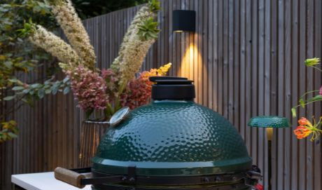 This image depicts a cozy and inviting outdoor cooking setup in a lush, garden-like environment. The focal point is a large, green egg-shaped charcoal grill or smoker, which is surrounded by a wooden fence and framed by various ornamental plants and flowers. The grill is positioned on a white, minimalist counter or table, and there are small tealight candles or lanterns providing soft, warm lighting. The overall impression is one of a tranquil and well-designed outdoor living space, ideal for entertaining or relaxing while preparing delicious meals.