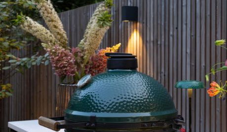 This image depicts a cozy and inviting outdoor cooking setup in a lush, garden-like environment. The focal point is a large, green egg-shaped charcoal grill or smoker, which is surrounded by a wooden fence and framed by various ornamental plants and flowers. The grill is positioned on a white, minimalist counter or table, and there are small tealight candles or lanterns providing soft, warm lighting. The overall impression is one of a tranquil and well-designed outdoor living space, ideal for entertaining or relaxing while preparing delicious meals.