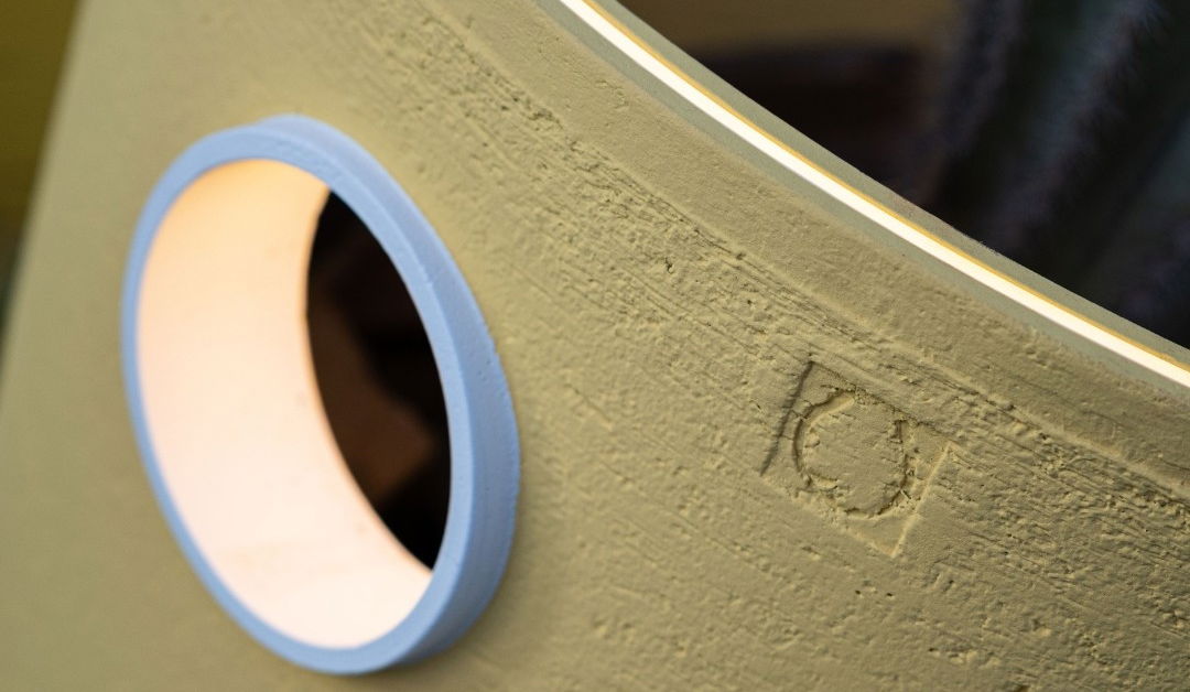 This image shows a close-up view of a textured, olive-green surface, likely a wall or furniture piece. The surface has a rough, almost brushed or sponged appearance, adding depth and character to the material. A round, light-colored shape can be seen partially in the frame, suggesting the presence of some kind of lighting fixture or decorative element integrated into the surface.  The image emphasizes the tactile and visually interesting qualities of the textured material, inviting the viewer to imagine the feel and overall aesthetic of the object or space it is a part of. The lighting or reflective element adds a subtle highlight, adding to the visual interest and depth of the surface.  Overall, this image captures the nuanced details and thoughtful design of a carefully crafted, textured material that likely plays a role in creating a distinctive and visually appealing interior or architectural environment.