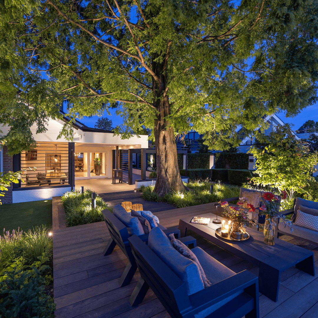 This image depicts a cozy and inviting outdoor living space nestled within a lush, verdant garden environment. The focal point is a comfortable seating area composed of plush blue sofas and armchairs, arranged around a low wooden coffee table.  The space is illuminated by a combination of warm, ambient lighting from wall-mounted fixtures and soft, diffused lighting from the overhead canopy structure. This creates a cozy and intimate atmosphere, complementing the natural textures and materials used throughout the design.  Surrounding the seating area, the garden is filled with a variety of vibrant, tropical foliage, including lush palm fronds and other verdant plants that frame the space and contribute to the serene, natural ambiance.  The overall composition emphasizes the integration of the built environment and the natural landscape, creating a harmonious and visually striking outdoor oasis that blends seamlessly with its surroundings. The combination of comfortable furnishings, thoughtful lighting, and the verdant, lush vegetation results in a captivating and inviting outdoor living space.