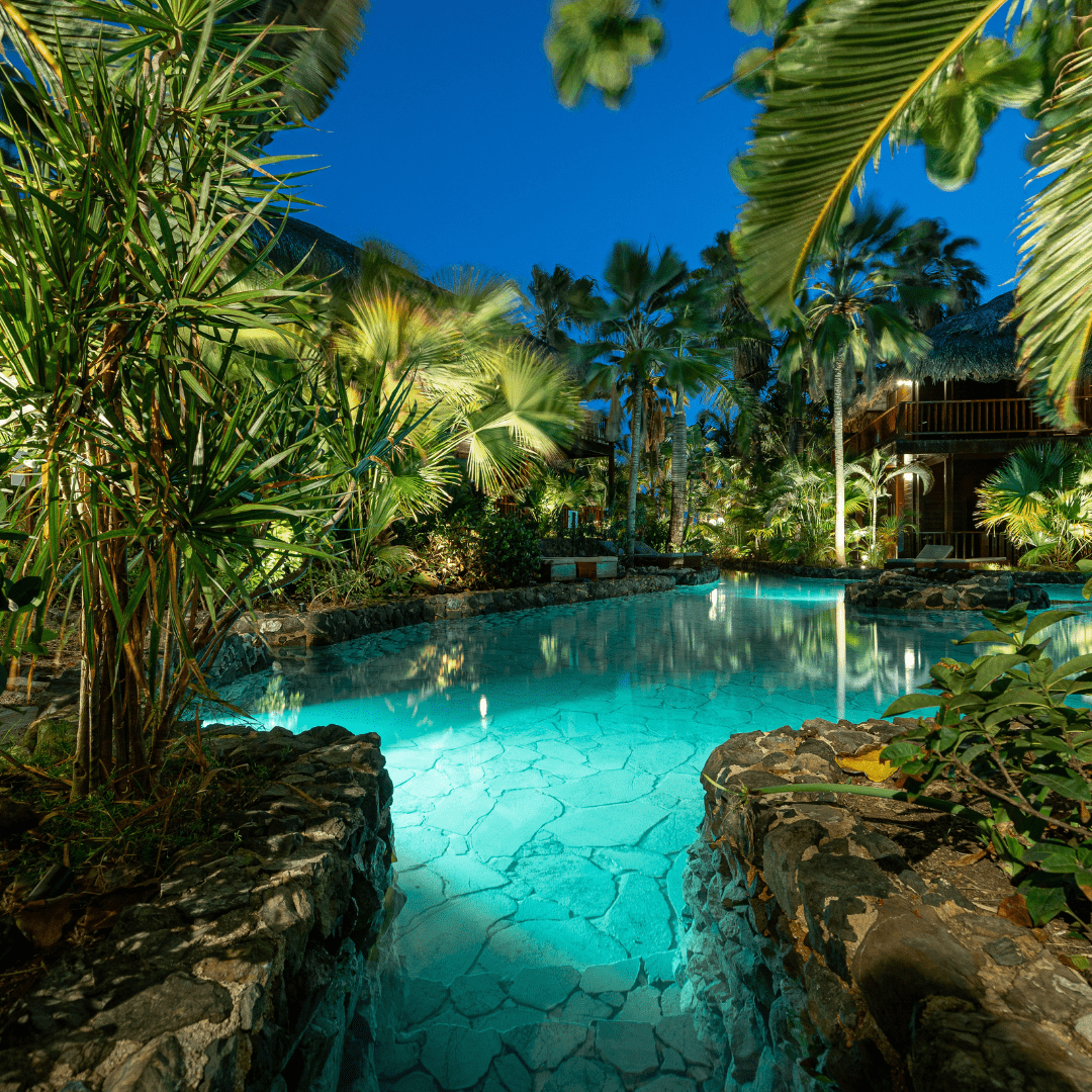 This image depicts a cozy and inviting outdoor living space nestled within a lush, verdant garden environment. The focal point is a comfortable seating area composed of plush blue sofas and armchairs, arranged around a low wooden coffee table.  The space is illuminated by a combination of warm, ambient lighting from wall-mounted fixtures and soft, diffused lighting from the overhead canopy structure. This creates a cozy and intimate atmosphere, complementing the natural textures and materials used throughout the design.  Surrounding the seating area, the garden is filled with a variety of vibrant, tropical foliage, including lush palm fronds and other verdant plants that frame the space and contribute to the serene, natural ambiance.  The overall composition emphasizes the integration of the built environment and the natural landscape, creating a harmonious and visually striking outdoor oasis that blends seamlessly with its surroundings. The combination of comfortable furnishings, thoughtful lighting, and the verdant, lush vegetation results in a captivating and inviting outdoor living space.