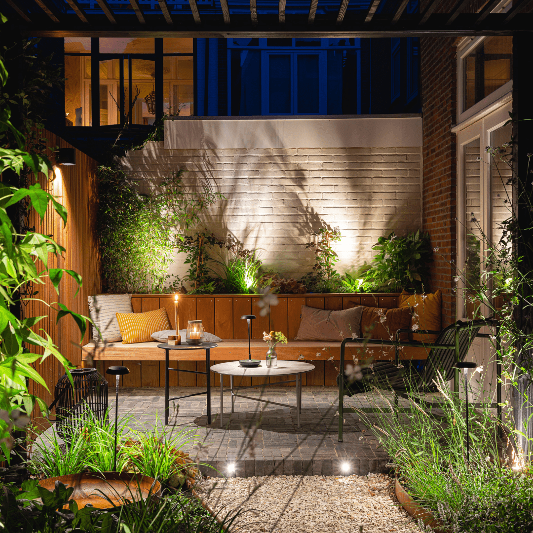This image depicts a cozy and inviting outdoor living space nestled within a lush, verdant garden environment. The focal point is a comfortable seating area composed of plush blue sofas and armchairs, arranged around a low wooden coffee table.  The space is illuminated by a combination of warm, ambient lighting from wall-mounted fixtures and soft, diffused lighting from the overhead canopy structure. This creates a cozy and intimate atmosphere, complementing the natural textures and materials used throughout the design.  Surrounding the seating area, the garden is filled with a variety of vibrant, tropical foliage, including lush palm fronds and other verdant plants that frame the space and contribute to the serene, natural ambiance.  The overall composition emphasizes the integration of the built environment and the natural landscape, creating a harmonious and visually striking outdoor oasis that blends seamlessly with its surroundings. The combination of comfortable furnishings, thoughtful lighting, and the verdant, lush vegetation results in a captivating and inviting outdoor living space.