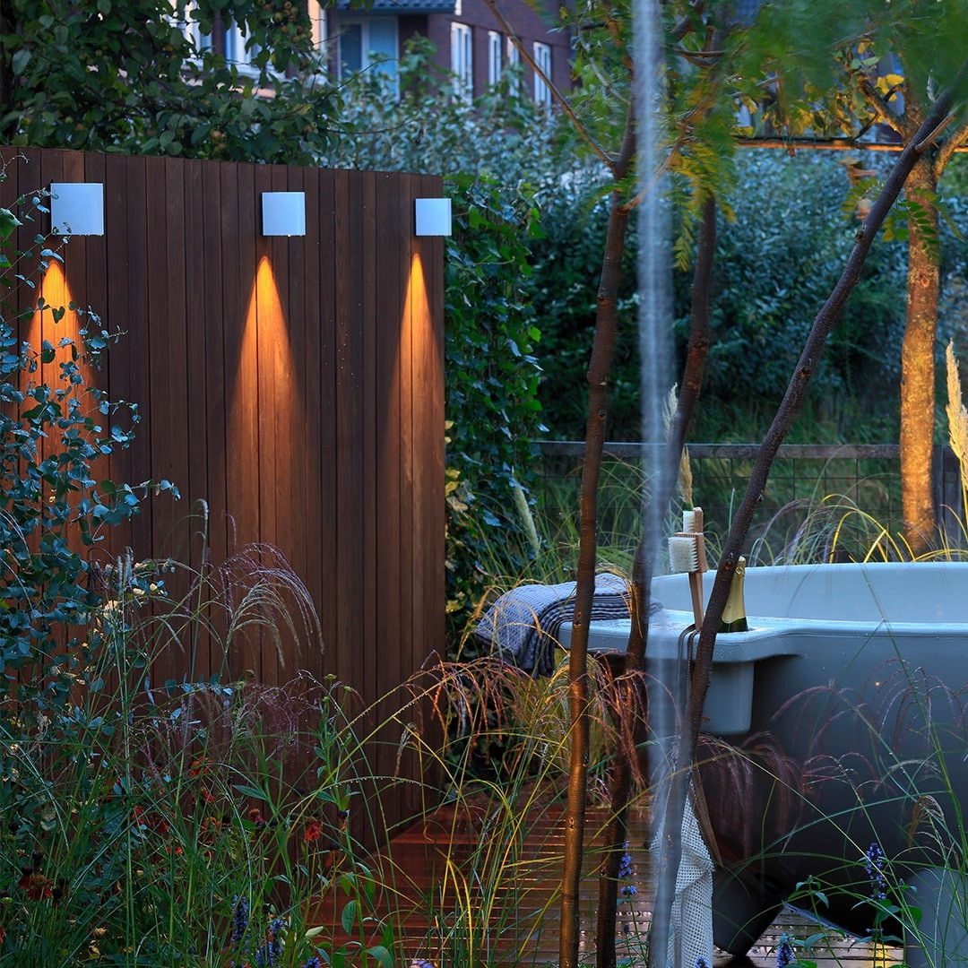 This image depicts a cozy and inviting outdoor living space, where the natural environment and the built elements seamlessly blend together to create a harmonious and visually appealing atmosphere. The focal point is the wooden fence, which serves as the backdrop for the scene. The fence is adorned with strategically placed lighting fixtures, whose warm, amber glow adds a sense of coziness and ambiance to the space. The lighting not only enhances the aesthetic appeal of the fence but also creates a welcoming and intimate ambiance as the evening approaches. Surrounding the fence, the lush, verdant foliage serves as a natural counterpoint to the built elements, creating a visually striking contrast. The varied textures and colors of the plants, ranging from the deep greens to the warm, golden hues, add depth and visual interest to the scene. In the foreground, a hot tub appears to be nestled within the natural setting, further enhancing the sense of relaxation and escape. The integration of the hot tub into the landscape, with the surrounding plants and the gentle waterfall, creates a serene and rejuvenating retreat. The overall design of this outdoor living space showcases a thoughtful and intentional approach to blending the built and natural elements, resulting in a harmonious and visually captivating environment. The attention to detail, from the lighting placement to the strategic use of plant materials, elevates the user's experience and creates a truly inviting and tranquil outdoor oasis.