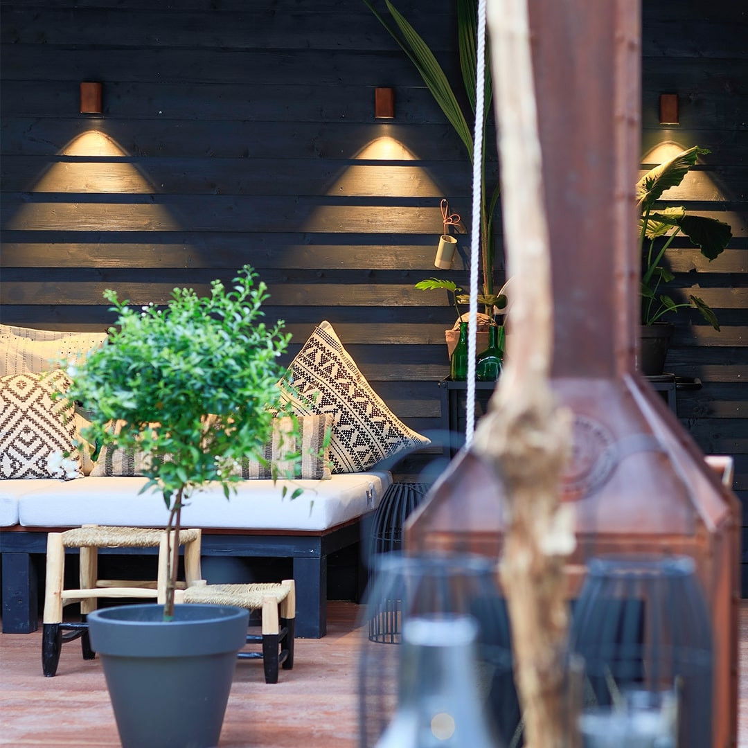 This image depicts a cozy and inviting outdoor living space, featuring a thoughtfully designed seating area with a mix of contemporary and rustic elements. The focal point is the comfortable bench seating, adorned with patterned cushions and accented by decorative throw pillows, creating a warm and welcoming atmosphere. The seating area is set against a backdrop of dark, wooden slat walls, which provide a striking contrast and a sense of enclosure to the space. Strategically placed lighting fixtures, with their warm, ambient glow, illuminate the area and contribute to the overall cozy ambiance. The greenery, including a potted plant and other natural elements, adds a touch of liveliness and softens the overall aesthetic. The inclusion of various decorative items, such as the patterned throw pillows and the glass bottle, suggests a curated and intentional approach to the space's design. The combination of natural, rustic, and contemporary elements creates a visually interesting and harmonious outdoor retreat. This image showcases the potential of a well-designed outdoor living space to provide a comfortable and inviting setting for relaxation, socializing, or simply enjoying the natural surroundings. The attention to detail and the thoughtful integration of various design elements contribute to the overall sense of cohesion and sophistication within this charming outdoor oasis.