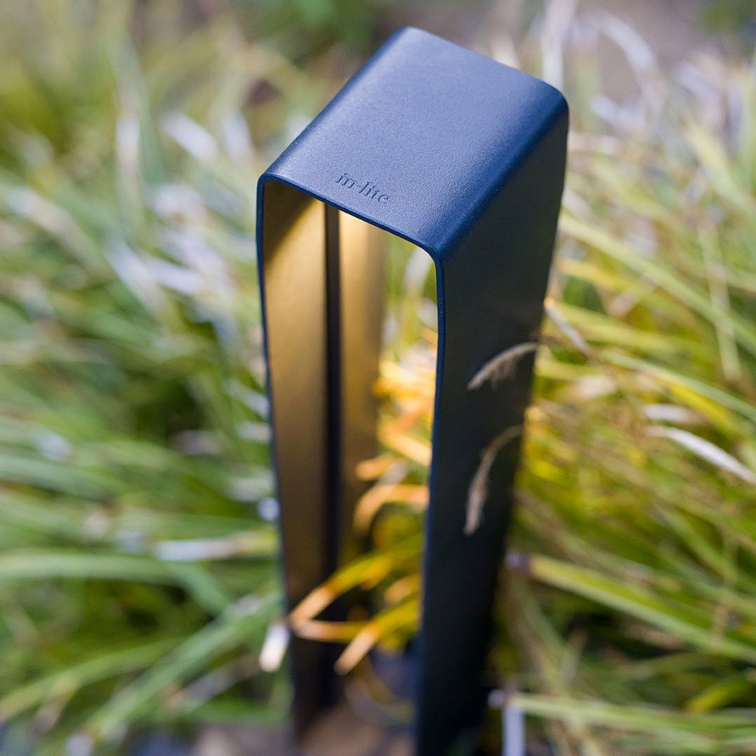 This image showcases a sleek and modern outdoor lighting fixture, seamlessly integrated into a lush, natural environment. The lighting fixture, with its clean, minimalist design and dark blue-gray finish, appears to be a product from the brand "flo-lite". The fixture is positioned amidst a vibrant, verdant backdrop of foliage, creating a visually striking contrast between the built and natural elements. The warm, diffused glow emitted by the lighting adds a sense of ambiance and coziness to the outdoor space, complementing the organic textures and colors of the surrounding vegetation. The strategic placement of the lighting fixture suggests a thoughtful approach to landscape design, where the built and natural elements are carefully balanced to create a cohesive and harmonious visual experience. The clean, contemporary aesthetic of the lighting fixture enhances the overall sophistication and refinement of the outdoor setting. This image demonstrates the importance of considered lighting design in elevating the aesthetic and functional qualities of an outdoor living space. The incorporation of this discrete, yet impactful lighting fixture allows the natural beauty of the surroundings to take center stage, while providing a warm and inviting ambiance for the user to enjoy.