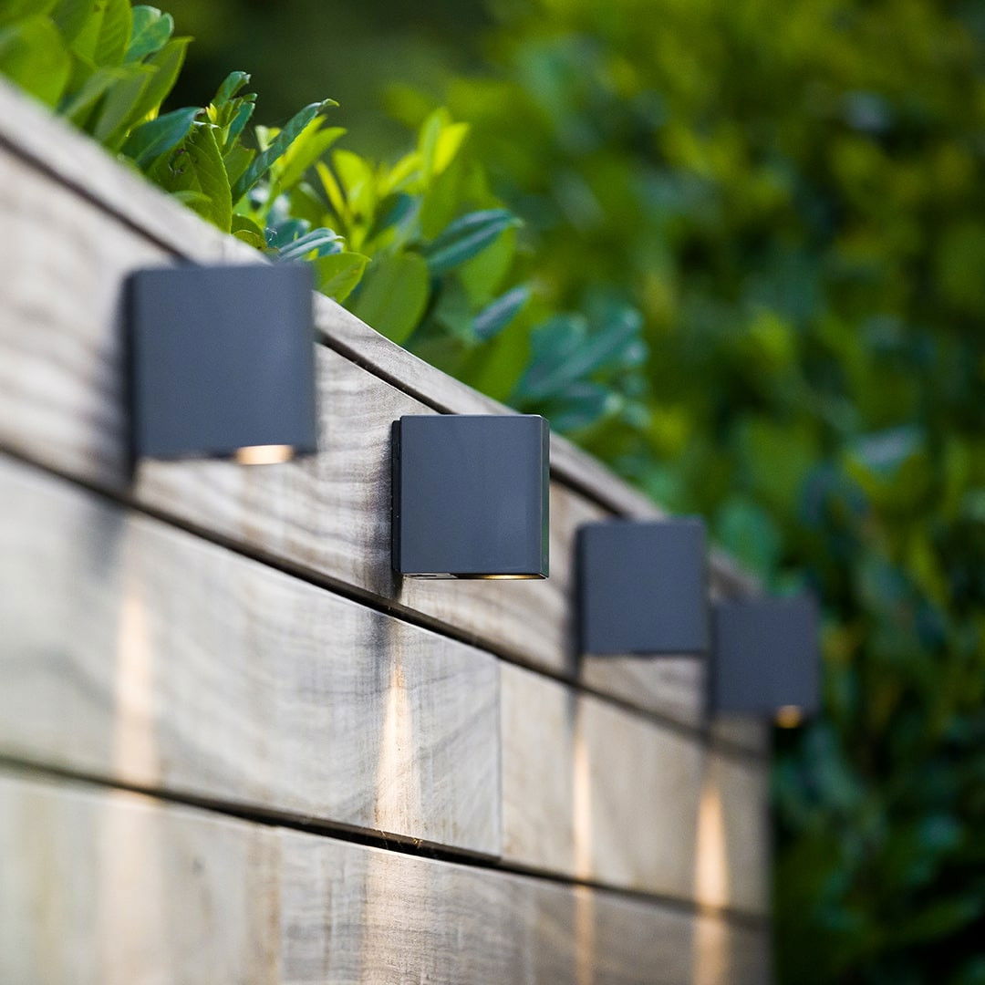 This image showcases a thoughtfully designed outdoor lighting solution, seamlessly integrated into a lush, natural environment. The lighting fixtures, with their sleek and minimalist black casing, are strategically positioned along the wooden fence, partially obscured by the vibrant greenery. The placement of the lighting creates a warm and inviting ambiance, complementing the organic textures and colors of the surrounding foliage. The warm, diffused glow emitted by the fixtures adds a sense of coziness and sophistication to the outdoor space, enhancing the overall aesthetic. The integration of the lighting into the wooden fence structure suggests a well-considered design approach, where the built and natural elements are harmoniously combined. The discreet placement of the fixtures allows the natural beauty of the environment to take center stage, while still providing the necessary illumination to create a welcoming and inviting atmosphere. This image exemplifies the power of thoughtful outdoor lighting design to elevate the overall experience and enjoyment of an outdoor living space. By seamlessly blending the artificial and natural elements, the designer has created a visually striking and functionally effective solution that seamlessly enhances the user's experience of the outdoor environment.