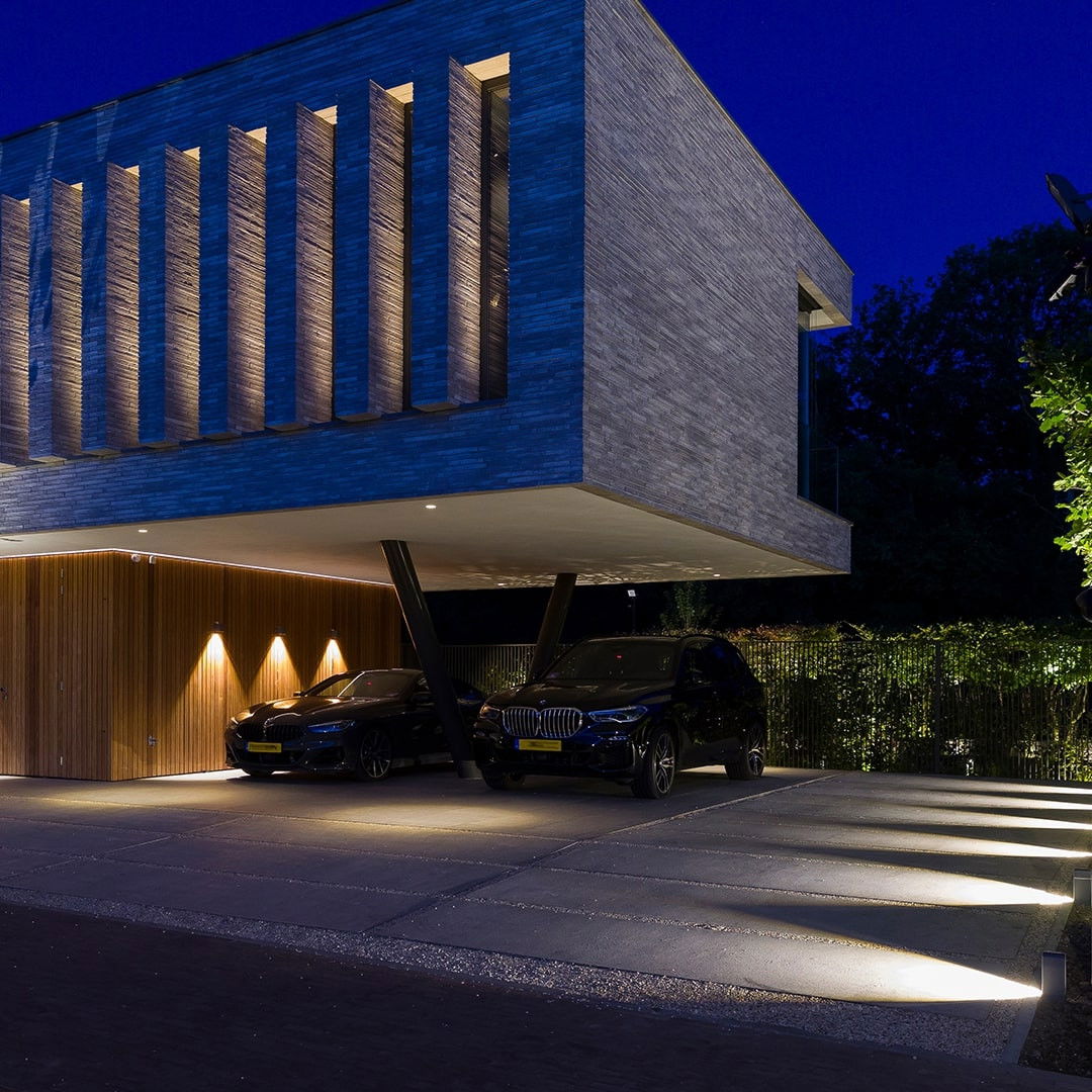 This striking image depicts a contemporary and sophisticated residential building, with a distinct architectural design that seamlessly blends modern elements and natural surroundings. The focal point is the building's facade, which features a bold, angular composition of vertical lines and a cantilevered overhang. The material palette includes a combination of warm, wooden elements and a cool, stone-like cladding that creates a visually intriguing contrast. The carefully placed lighting, with its warm, diffused glow, illuminates the building's exterior and accentuates the architectural details, casting dramatic shadows and highlighting the interplay of light and shadow. This lighting design not only enhances the aesthetic appeal of the structure but also contributes to a welcoming and inviting atmosphere. Two high-end vehicles, presumably belonging to the residents, are parked in the foreground, adding a touch of sophistication and exclusivity to the scene. The smooth, paved surface and the surrounding lush greenery further complement the overall design, creating a harmonious integration of the built and natural environments. This image showcases the attention to detail and the thoughtful approach to architectural design that can elevate a residential property, transforming it into a visually striking and immersive experience. The combination of bold, contemporary forms, carefully curated materials, and strategic lighting design results in a captivating and modern residential space that seamlessly blends with its natural surroundings.