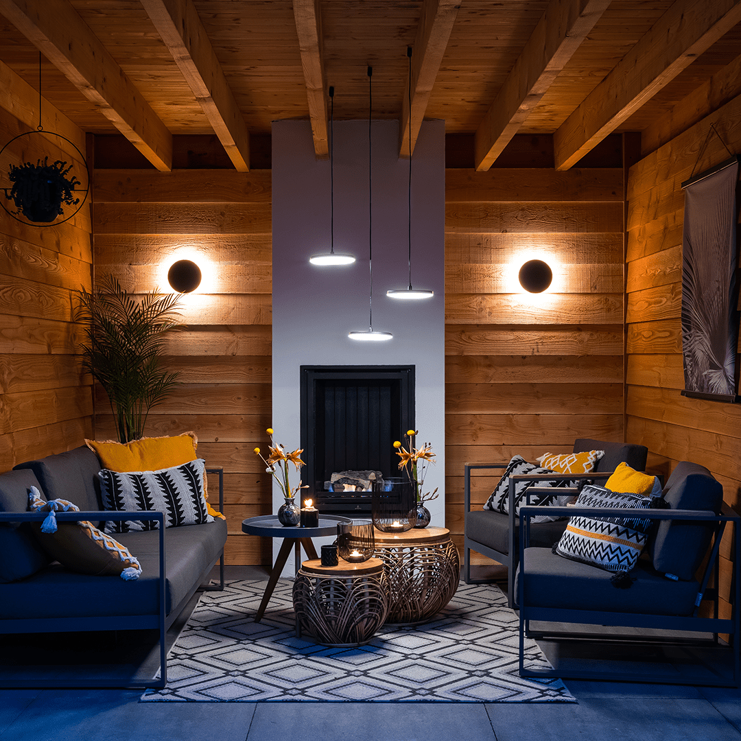 This image depicts a cozy and inviting outdoor living space nestled within a lush, verdant garden environment. The focal point is a comfortable seating area composed of plush blue sofas and armchairs, arranged around a low wooden coffee table.  The space is illuminated by a combination of warm, ambient lighting from wall-mounted fixtures and soft, diffused lighting from the overhead canopy structure. This creates a cozy and intimate atmosphere, complementing the natural textures and materials used throughout the design.  Surrounding the seating area, the garden is filled with a variety of vibrant, tropical foliage, including lush palm fronds and other verdant plants that frame the space and contribute to the serene, natural ambiance.  The overall composition emphasizes the integration of the built environment and the natural landscape, creating a harmonious and visually striking outdoor oasis that blends seamlessly with its surroundings. The combination of comfortable furnishings, thoughtful lighting, and the verdant, lush vegetation results in a captivating and inviting outdoor living space.