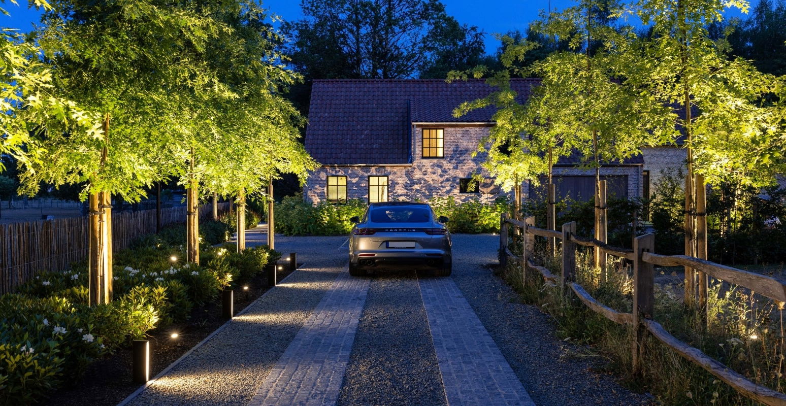 This image depicts a beautifully landscaped and illuminated outdoor space surrounding a stone and brick residence. The main focal point is the driveway leading up to the house, which is paved with a combination of gravel and brick, creating a visually appealing textural contrast. The driveway is flanked by lush, well-maintained landscaping, including mature trees with vibrant foliage that is highlighted by strategically placed outdoor lighting. These lighting fixtures create a warm, ambient glow, enhancing the natural beauty of the surrounding greenery and the architectural features of the home. The residence itself is a charming, historic-looking building with a stone facade and a tiled roof, blending seamlessly with the natural setting. The windows are illuminated, suggesting a cozy and inviting interior that complements the tranquil, serene atmosphere of the outdoor space. Overall, this image conveys a sense of refined elegance and a harmonious integration of the built environment and the natural landscape. The carefully considered lighting design, the meticulously landscaped grounds, and the architectural character of the home all contribute to the creation of a visually striking and inviting outdoor living space.