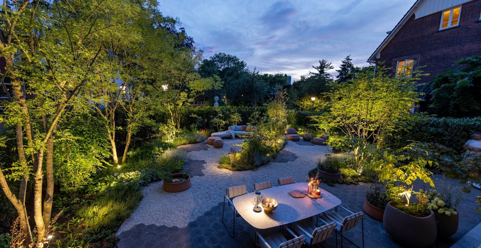 This image depicts a beautifully designed and meticulously landscaped outdoor living and entertainment space, centered around a serene and inviting garden setting. The scene is illuminated by a combination of ambient lighting, creating a warm and enchanting atmosphere as the sun sets. The focal point of the image is the tranquil garden area, featuring a winding pathway made of gravel and stone that leads through the lush, well-maintained greenery. The path is flanked by various seating areas, including a stylish dining table set with lanterns and accessories, as well as comfortable lounge furniture nestled among the foliage. The landscaping is a highlight, with a variety of mature trees, shrubs, and other plants that create a lush, natural canopy overhead. Strategically placed lighting fixtures illuminate the pathway and the various seating areas, casting a warm glow that accentuates the textures and colors of the plants and hardscaping. In the background, the warm, illuminated windows of the home suggest a cozy and inviting indoor space that complements the tranquil outdoor setting. The overall composition emphasizes the harmonious integration of the built environment and the natural elements, resulting in a visually striking and serene outdoor oasis that is perfect for both relaxation and entertainment. The attention to detail, from the thoughtful lighting design to the meticulously maintained greenery, contributes to the sophisticated and elegant character of the space. This image conveys a sense of refined luxury, comfort, and an appreciation for the beauty of the outdoors, where the designed and the natural seamlessly coexist to create a captivating and inviting retreat.