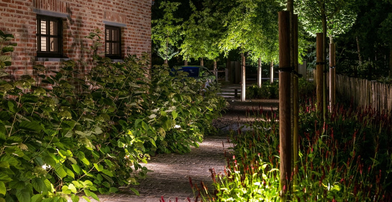 This image depicts a beautifully landscaped and illuminated outdoor pathway leading through a lush, natural garden setting. The scene is centered around a winding gravel path that winds through a verdant, densely planted area, creating a serene and inviting atmosphere. The path is flanked by a variety of colorful and textural plants, including tall grasses, vibrant foliage, and flowering plants that add visual interest and depth to the space. Strategically placed lighting fixtures along the path cast a warm, ambient glow, highlighting the rich textures and colors of the vegetation. In the background, a historic-looking brick building with illuminated windows suggests a cozy and inviting indoor space that complements the tranquil outdoor setting. The overall composition emphasizes the harmonious integration of the built environment and the natural elements, creating a visually striking and serene outdoor retreat. The attention to detail in the landscaping and lighting design contributes to the sophisticated and elegant character of the space. This image conveys a sense of refined luxury, comfort, and an appreciation for the beauty of the natural world, where the designed and the wild seamlessly coexist to create a captivating and inviting outdoor oasis.