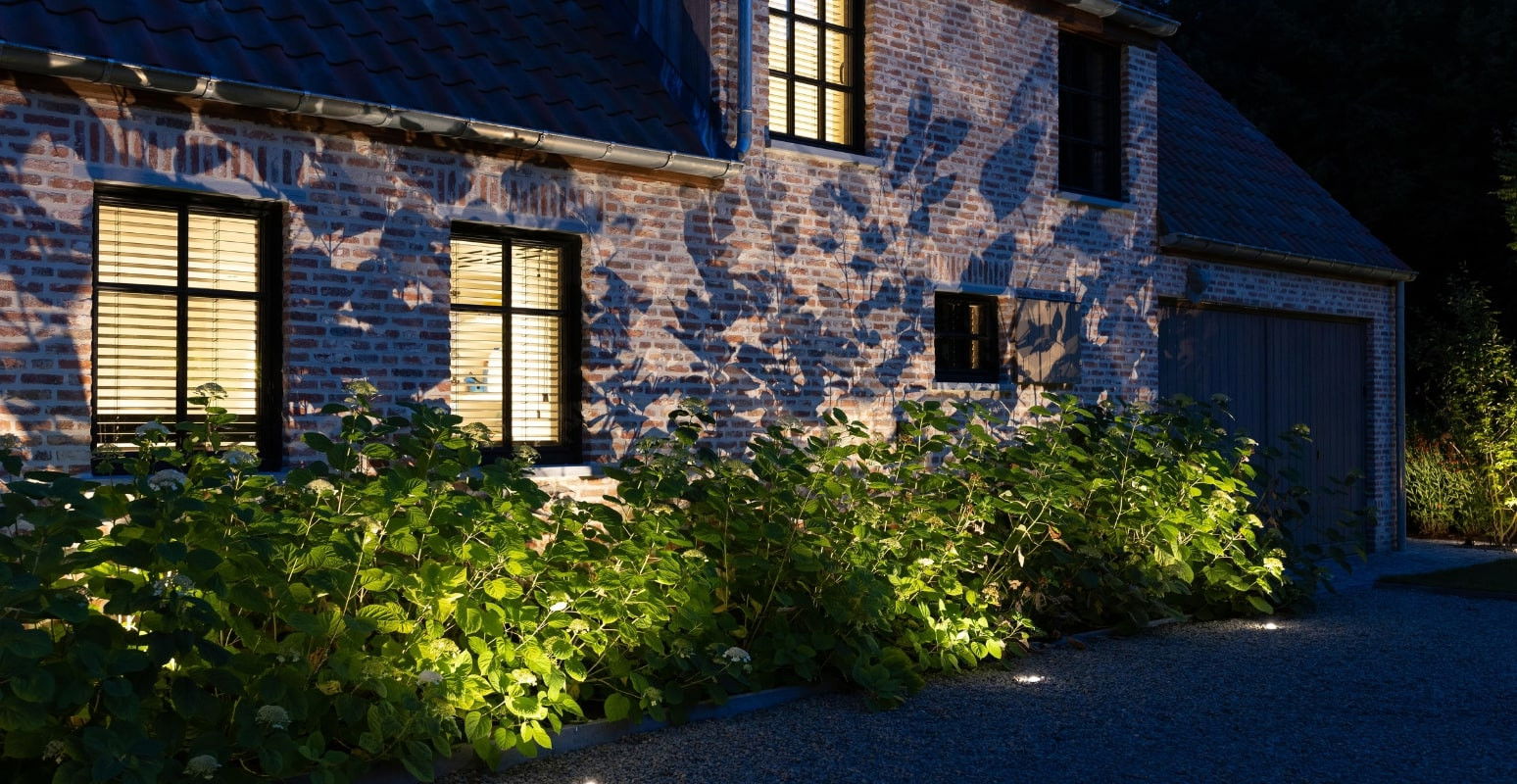 This image depicts a charming and cozy outdoor scene, centered around a historic brick building with illuminated windows and a lush, densely planted garden surrounding it. The building's facade is highlighted by strategic lighting, casting a warm, inviting glow that contrasts with the cool, blue-tinted shadows of the night. The brick walls, with their distinctive texture and patterns, add visual interest and a sense of timeless character to the scene. Surrounding the building is a vibrant, well-maintained garden, with a variety of plants and foliage spilling out onto the path and creating a sense of enclosure and intimacy. The rich, verdant greens of the plants are accentuated by the targeted lighting, which casts dramatic shadows and highlights the natural textures and shapes of the vegetation. The overall composition creates a serene and enchanting atmosphere, where the historic architecture and the lush, naturalistic landscaping coexist in a harmonious and visually striking way. The attention to detail in the lighting design and the careful curation of the garden elements contribute to the sense of refined elegance and a deep appreciation for the beauty of the natural world. This image invites the viewer to imagine stepping into this cozy, inviting outdoor oasis, where the built environment and the natural landscape seamlessly blend together to create a captivating and tranquil retreat.