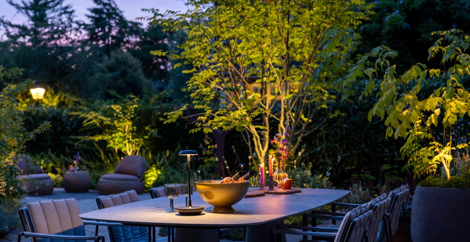 This image depicts a beautifully designed and illuminated outdoor dining space, set against a lush and vibrant natural landscape. The focal point is the striking wooden dining table, which is surrounded by comfortable-looking chairs and accented with an array of decorative elements, including a large golden-colored serving bowl and various other tableware. The table and its setting are positioned in a way that takes full advantage of the stunning natural surroundings. Overhead, the trees are illuminated with warm, subtle lighting, creating a cozy and inviting ambiance. The vibrant greens and yellows of the foliage contrast beautifully with the deep blues and purples of the evening sky, resulting in a visually striking and harmonious scene. The attention to detail in the lighting design, the careful curation of the table setting, and the integration of the natural elements all contribute to the overall sense of refinement and elegance. This outdoor dining area appears to be a thoughtfully designed and meticulously maintained space, where one could imagine enjoying a peaceful and memorable meal in the company of others. The overall composition conveys a sense of tranquility, comfort, and a deep appreciation for the beauty of the natural world. The warm lighting, the rich textures of the wood and foliage, and the inviting atmosphere all work together to create an enchanting and captivating outdoor retreat.