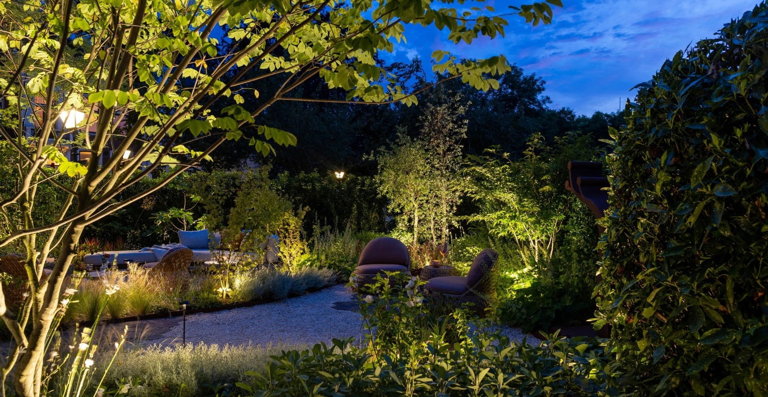 This image depicts a beautifully designed and meticulously maintained outdoor living space, bathed in the warm glow of strategically placed lighting. The scene is centered around a winding gravel pathway that leads through a lush, verdant garden, surrounded by a variety of mature trees and shrubs. The path is flanked by comfortable, inviting seating areas, including plush lounge chairs and benches, all nestled amongst the rich foliage and carefully curated plantings. The lighting fixtures, which are situated along the pathway and throughout the garden, cast a soft, ambient illumination that highlights the natural textures and colors of the plants and hardscaping. In the background, the deep blue of the evening sky provides a dramatic contrast to the vibrant greens and yellows of the foliage, creating a sense of depth and visual interest. The overall composition emphasizes the harmonious integration of the designed elements and the natural landscape, resulting in a serene and captivating outdoor oasis. The attention to detail, from the thoughtful placement of the lighting to the well-maintained plantings, contributes to the sophisticated and elegant character of the space. This image conveys a sense of refined luxury, comfort, and a deep appreciation for the beauty of the natural world, where the designed and the wild seamlessly coexist to create a captivating and inviting retreat.