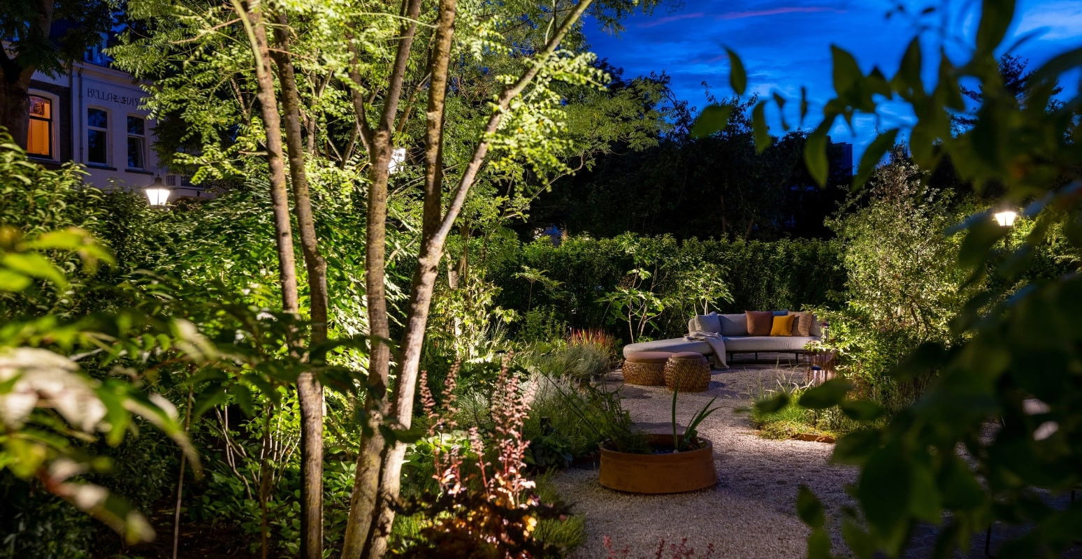 This image depicts a beautifully designed and meticulously maintained outdoor living space, bathed in the warm glow of strategically placed lighting. The scene is centered around a winding gravel pathway that leads through a lush, verdant garden, surrounded by a variety of mature trees and shrubs. The path is flanked by comfortable, inviting seating areas, including plush lounge chairs and benches, all nestled amongst the rich foliage and carefully curated plantings. The lighting fixtures, which are situated along the pathway and throughout the garden, cast a soft, ambient illumination that highlights the natural textures and colors of the plants and hardscaping. In the background, the deep blue of the evening sky provides a dramatic contrast to the vibrant greens and yellows of the foliage, creating a sense of depth and visual interest. The overall composition emphasizes the harmonious integration of the designed elements and the natural landscape, resulting in a serene and captivating outdoor oasis. The attention to detail, from the thoughtful placement of the lighting to the well-maintained plantings, contributes to the sophisticated and elegant character of the space. This image conveys a sense of refined luxury, comfort, and a deep appreciation for the beauty of the natural world, where the designed and the wild seamlessly coexist to create a captivating and inviting retreat.