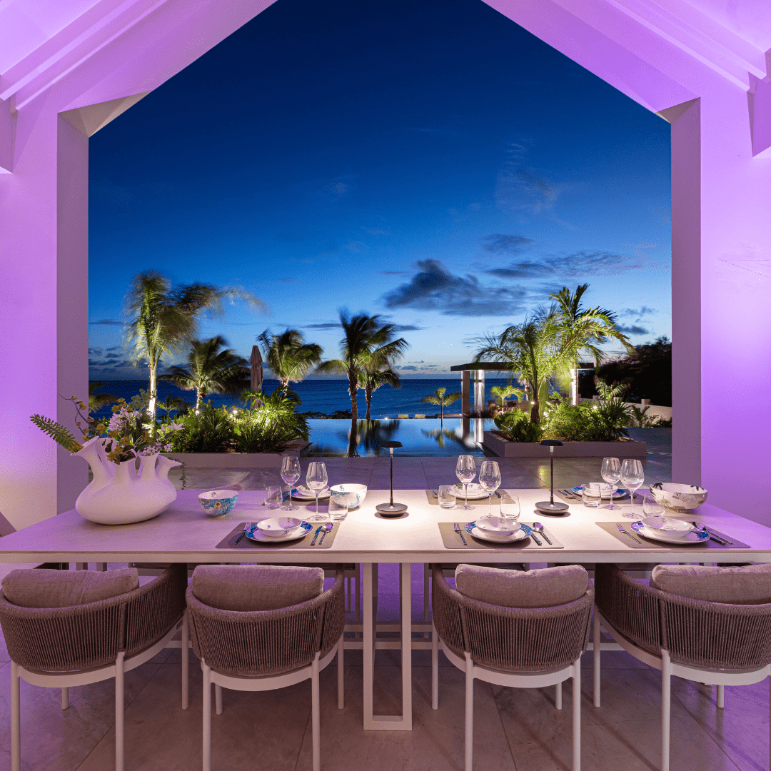 This image depicts a cozy and inviting outdoor living space nestled within a lush, verdant garden environment. The focal point is a comfortable seating area composed of plush blue sofas and armchairs, arranged around a low wooden coffee table. The space is illuminated by a combination of warm, ambient lighting from wall-mounted fixtures and soft, diffused lighting from the overhead canopy structure. This creates a cozy and intimate atmosphere, complementing the natural textures and materials used throughout the design. Surrounding the seating area, the garden is filled with a variety of vibrant, tropical foliage, including lush palm fronds and other verdant plants that frame the space and contribute to the serene, natural ambiance. The overall composition emphasizes the integration of the built environment and the natural landscape, creating a harmonious and visually striking outdoor oasis that blends seamlessly with its surroundings. The combination of comfortable furnishings, thoughtful lighting, and the verdant, lush vegetation results in a captivating and inviting outdoor living space.