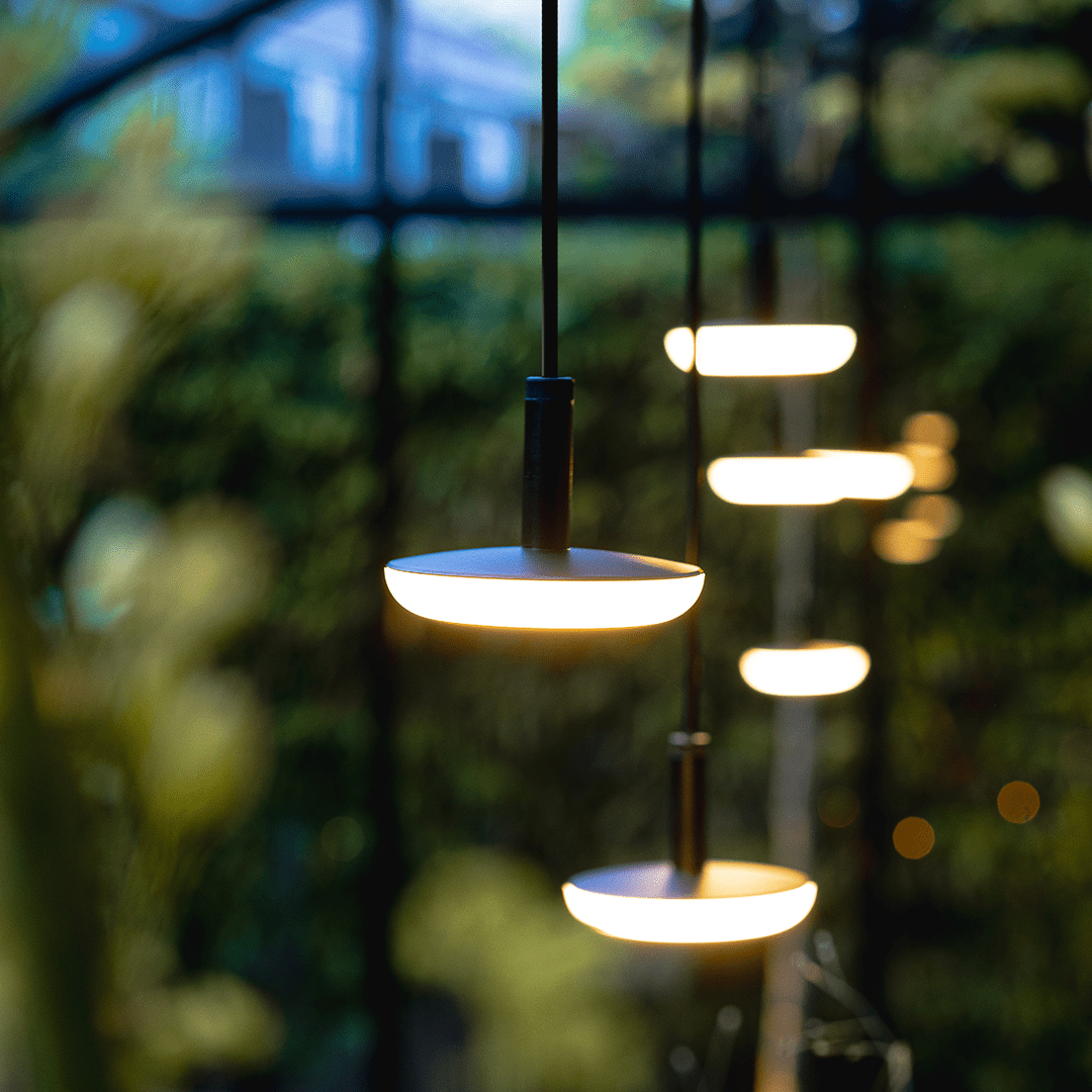 This image depicts a cozy and inviting outdoor living space nestled within a lush, verdant garden environment. The focal point is a comfortable seating area composed of plush blue sofas and armchairs, arranged around a low wooden coffee table.  The space is illuminated by a combination of warm, ambient lighting from wall-mounted fixtures and soft, diffused lighting from the overhead canopy structure. This creates a cozy and intimate atmosphere, complementing the natural textures and materials used throughout the design.  Surrounding the seating area, the garden is filled with a variety of vibrant, tropical foliage, including lush palm fronds and other verdant plants that frame the space and contribute to the serene, natural ambiance.  The overall composition emphasizes the integration of the built environment and the natural landscape, creating a harmonious and visually striking outdoor oasis that blends seamlessly with its surroundings. The combination of comfortable furnishings, thoughtful lighting, and the verdant, lush vegetation results in a captivating and inviting outdoor living space.
