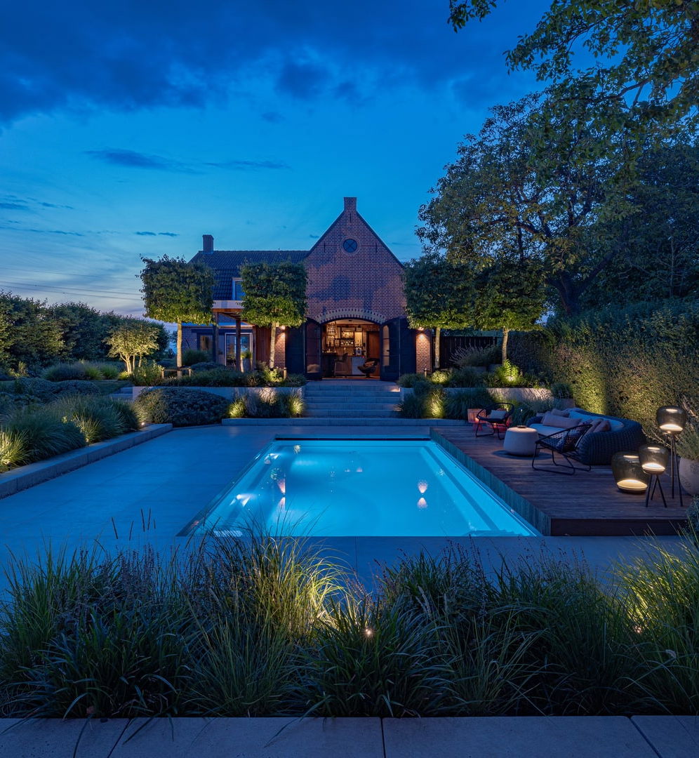 This image depicts a beautifully designed and meticulously landscaped outdoor living and entertainment space, centered around an inviting swimming pool. The scene is illuminated by a combination of subtle, ambient lighting, creating a warm and enchanting atmosphere. The focal point of the image is the rectangular swimming pool with its smooth, reflective surface that mirrors the surrounding landscape. The pool is surrounded by a wooden deck and interspersed with various seating areas, including a long table with chairs arranged for dining or socializing. The landscaping features an array of carefully curated plants, trees, and shrubbery, creating a natural, serene ambiance. Decorative lighting elements, such as wall-mounted fixtures and subtle pathway illumination, further enhance the overall atmosphere and highlight the architectural and landscape features. In the background, a charming brick building with architectural details can be seen, adding a historic and elegant element to the scene. The building is illuminated, creating a warm and inviting glow that complements the lighting around the pool. The overall composition emphasizes the harmonious integration of the built environment and the natural elements, resulting in a visually striking and inviting outdoor oasis that is perfect for both relaxation and entertainment. The attention to detail, from the thoughtful lighting design to the meticulously maintained greenery, contributes to the sophisticated and elegant character of the space. This image conveys a sense of refined luxury, comfort, and an appreciation for the beauty of the outdoors, where the designed and the natural seamlessly coexist to create a captivating and serene outdoor retreat.