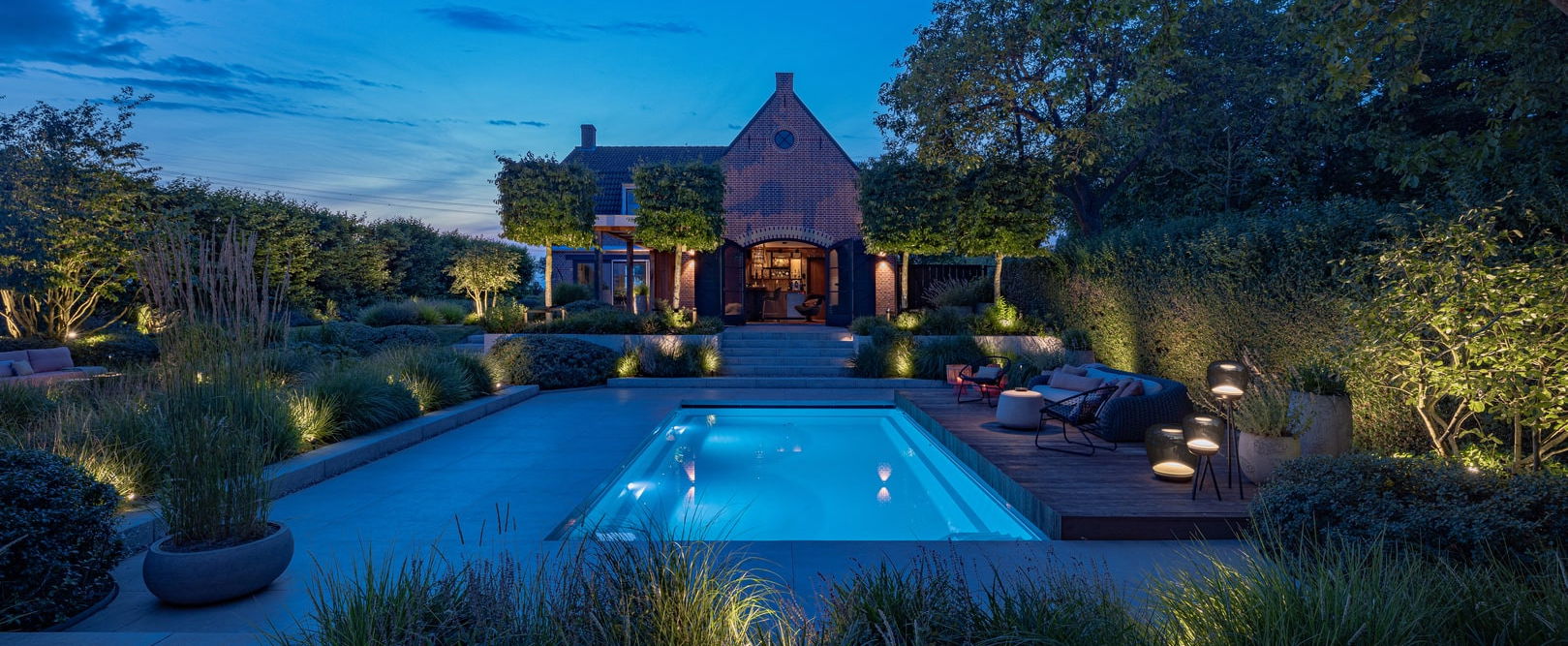 This image depicts a beautifully designed and meticulously landscaped outdoor living and entertainment space, centered around an inviting swimming pool. The scene is illuminated by a combination of subtle, ambient lighting, creating a warm and enchanting atmosphere. The focal point of the image is the rectangular swimming pool with its smooth, reflective surface that mirrors the surrounding landscape. The pool is surrounded by a wooden deck and interspersed with various seating areas, including a long table with chairs arranged for dining or socializing. The landscaping features an array of carefully curated plants, trees, and shrubbery, creating a natural, serene ambiance. Decorative lighting elements, such as wall-mounted fixtures and subtle pathway illumination, further enhance the overall atmosphere and highlight the architectural and landscape features. In the background, a charming brick building with architectural details can be seen, adding a historic and elegant element to the scene. The building is illuminated, creating a warm and inviting glow that complements the lighting around the pool. The overall composition emphasizes the harmonious integration of the built environment and the natural elements, resulting in a visually striking and inviting outdoor oasis that is perfect for both relaxation and entertainment. The attention to detail, from the thoughtful lighting design to the meticulously maintained greenery, contributes to the sophisticated and elegant character of the space. This image conveys a sense of refined luxury, comfort, and an appreciation for the beauty of the outdoors, where the designed and the natural seamlessly coexist to create a captivating and serene outdoor retreat.