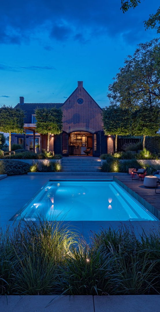 This image depicts a beautifully designed and meticulously landscaped outdoor living and entertainment space, centered around an inviting swimming pool. The scene is illuminated by a combination of subtle, ambient lighting, creating a warm and enchanting atmosphere. The focal point of the image is the rectangular swimming pool with its smooth, reflective surface that mirrors the surrounding landscape. The pool is surrounded by a wooden deck and interspersed with various seating areas, including a long table with chairs arranged for dining or socializing. The landscaping features an array of carefully curated plants, trees, and shrubbery, creating a natural, serene ambiance. Decorative lighting elements, such as wall-mounted fixtures and subtle pathway illumination, further enhance the overall atmosphere and highlight the architectural and landscape features. In the background, a charming brick building with architectural details can be seen, adding a historic and elegant element to the scene. The building is illuminated, creating a warm and inviting glow that complements the lighting around the pool. The overall composition emphasizes the harmonious integration of the built environment and the natural elements, resulting in a visually striking and inviting outdoor oasis that is perfect for both relaxation and entertainment. The attention to detail, from the thoughtful lighting design to the meticulously maintained greenery, contributes to the sophisticated and elegant character of the space. This image conveys a sense of refined luxury, comfort, and an appreciation for the beauty of the outdoors, where the designed and the natural seamlessly coexist to create a captivating and serene outdoor retreat.