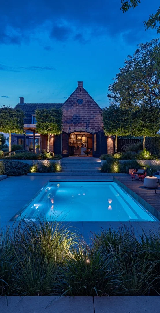 This image depicts a beautifully designed and meticulously landscaped outdoor living and entertainment space, centered around an inviting swimming pool. The scene is illuminated by a combination of subtle, ambient lighting, creating a warm and enchanting atmosphere. The focal point of the image is the rectangular swimming pool with its smooth, reflective surface that mirrors the surrounding landscape. The pool is surrounded by a wooden deck and interspersed with various seating areas, including a long table with chairs arranged for dining or socializing. The landscaping features an array of carefully curated plants, trees, and shrubbery, creating a natural, serene ambiance. Decorative lighting elements, such as wall-mounted fixtures and subtle pathway illumination, further enhance the overall atmosphere and highlight the architectural and landscape features. In the background, a charming brick building with architectural details can be seen, adding a historic and elegant element to the scene. The building is illuminated, creating a warm and inviting glow that complements the lighting around the pool. The overall composition emphasizes the harmonious integration of the built environment and the natural elements, resulting in a visually striking and inviting outdoor oasis that is perfect for both relaxation and entertainment. The attention to detail, from the thoughtful lighting design to the meticulously maintained greenery, contributes to the sophisticated and elegant character of the space. This image conveys a sense of refined luxury, comfort, and an appreciation for the beauty of the outdoors, where the designed and the natural seamlessly coexist to create a captivating and serene outdoor retreat.