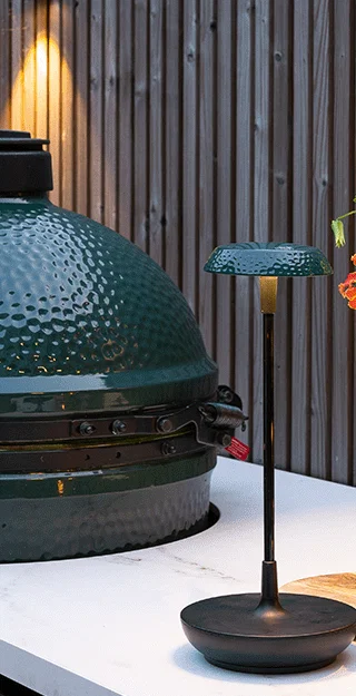 The image depicts a beautifully designed outdoor garden scene at night. The focal point is a large, decorative planter or urn filled with lush, vibrant foliage that is illuminated by strategic landscape lighting, creating a warm and inviting ambiance. Surrounding the planter are carefully placed stepping stones that lead to a covered patio or entryway visible in the background. The garden is further enhanced by the presence of a tall, tree-like plant with brilliant yellow leaves that add a striking contrast to the darker tones of the surrounding plants and hardscaping. The overall impression is one of a thoughtfully curated and meticulously maintained outdoor living space, blending natural elements with contemporary design. The strategic use of lighting, the harmonious integration of plants and materials, and the attention to detail create a visually captivating and serene nighttime garden oasis, inviting the viewer to immerse themselves in the tranquil and inviting atmosphere.