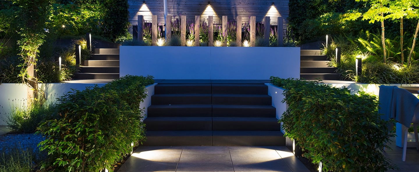 This image depicts a beautifully designed outdoor living space, featuring a raised seating area with a striking blue backdrop. The seating area is surrounded by lush, vibrant greenery, including tall grasses and other plant life that add texture and depth to the scene. The stairs leading up to the seating area are illuminated by strategically placed lighting fixtures, creating a warm and inviting atmosphere. The blue backdrop, which appears to be a wall or structure, adds a bold and modern touch to the overall design. The lighting throughout the space is used effectively, highlighting the architectural elements and the surrounding vegetation. The result is a harmonious and visually captivating outdoor retreat, where one could imagine spending time relaxing or entertaining guests. The attention to detail, the integration of the natural and built elements, and the overall design aesthetic create a sense of sophistication and elegance. This image invites the viewer to imagine themselves immersed in this tranquil and inviting outdoor oasis.