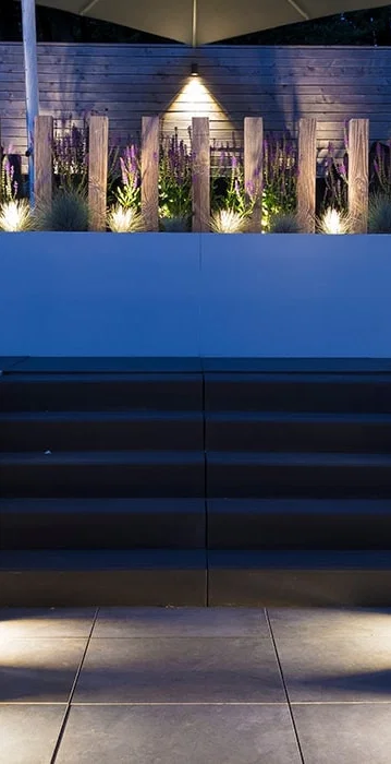 This image depicts a beautifully designed outdoor living space, featuring a raised seating area with a striking blue backdrop. The seating area is surrounded by lush, vibrant greenery, including tall grasses and other plant life that add texture and depth to the scene. The stairs leading up to the seating area are illuminated by strategically placed lighting fixtures, creating a warm and inviting atmosphere. The blue backdrop, which appears to be a wall or structure, adds a bold and modern touch to the overall design. The lighting throughout the space is used effectively, highlighting the architectural elements and the surrounding vegetation. The result is a harmonious and visually captivating outdoor retreat, where one could imagine spending time relaxing or entertaining guests. The attention to detail, the integration of the natural and built elements, and the overall design aesthetic create a sense of sophistication and elegance. This image invites the viewer to imagine themselves immersed in this tranquil and inviting outdoor oasis.