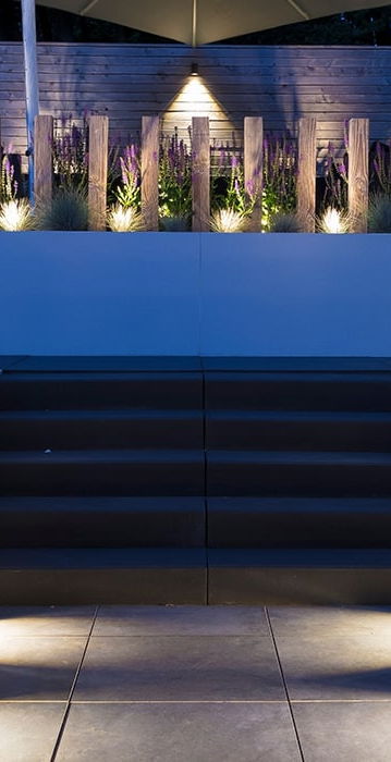 This image depicts a beautifully designed outdoor living space, featuring a raised seating area with a striking blue backdrop. The seating area is surrounded by lush, vibrant greenery, including tall grasses and other plant life that add texture and depth to the scene. The stairs leading up to the seating area are illuminated by strategically placed lighting fixtures, creating a warm and inviting atmosphere. The blue backdrop, which appears to be a wall or structure, adds a bold and modern touch to the overall design. The lighting throughout the space is used effectively, highlighting the architectural elements and the surrounding vegetation. The result is a harmonious and visually captivating outdoor retreat, where one could imagine spending time relaxing or entertaining guests. The attention to detail, the integration of the natural and built elements, and the overall design aesthetic create a sense of sophistication and elegance. This image invites the viewer to imagine themselves immersed in this tranquil and inviting outdoor oasis.