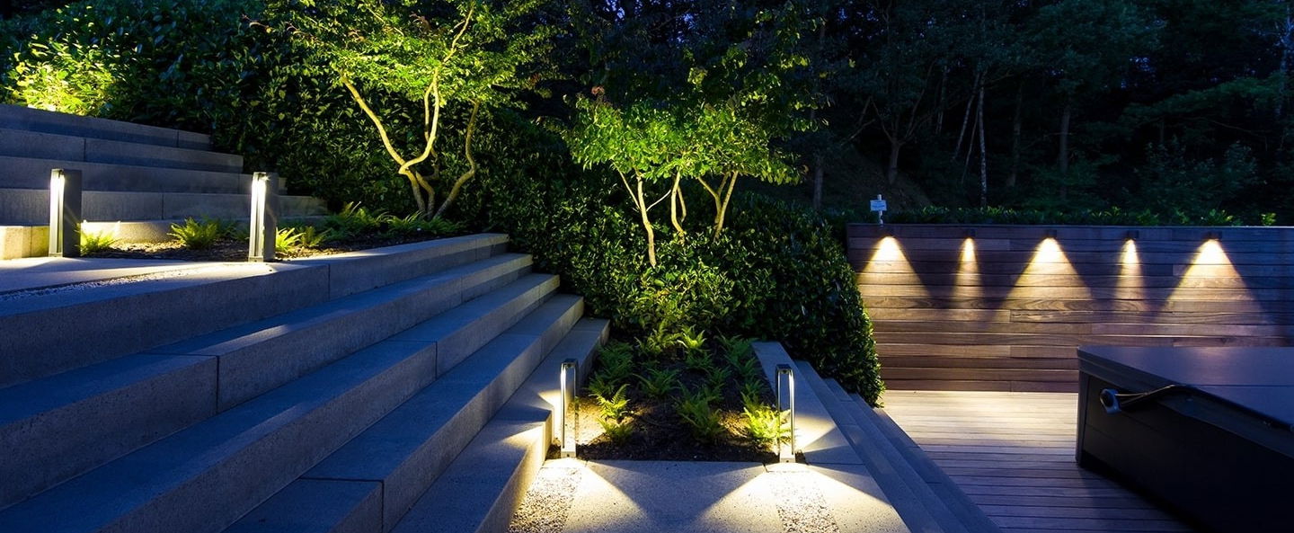 This image presents a stunning and well-designed outdoor living space that seamlessly integrates architectural elements with the surrounding natural environment. The focal point is the series of concrete steps leading down into the garden area, which are illuminated by strategically placed lighting fixtures that cast a warm and inviting glow. The steps themselves are intentionally designed with a modern, minimalist aesthetic, creating a clean and sleek visual contrast against the lush, verdant foliage that cascades over the edges. Flanking the steps, a wooden deck or platform extends the living space, further enhancing the connection between the built and natural elements. The warm wood tones of the deck complement the cool, gray concrete, while the lighting fixtures along the deck's edge add a sense of depth and drama to the overall composition. The carefully curated planting palette, with its vibrant greens and sculptural forms, creates a captivating backdrop that softens the architectural lines and adds a sense of natural harmony. The strategic placement of the lighting not only illuminates the steps and deck but also accentuates the texture and depth of the vegetation, casting dramatic shadows and highlighting the interplay of light and shadow. The integration of these various design elements – the concrete, wood, lighting, and lush greenery – results in a cohesive and visually striking outdoor living space that invites the user to engage with and explore the carefully crafted environment. This image exemplifies the power of thoughtful landscape and architectural design to create an immersive and harmonious outdoor experience.