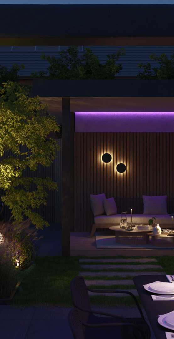 The image depicts a beautifully designed outdoor living space at night. The focal point is a covered seating area with plush, purple-colored sofas and chairs, creating a cozy and inviting atmosphere. This seating area is illuminated by subtle, warm lighting, both overhead and mounted on the surrounding wooden walls. The outdoor space is further enhanced by the presence of a lush, mature tree with vibrant green foliage, which adds a natural and serene element to the scene. A dining table set with dinnerware is also visible, suggesting this area is designed for both relaxation and al fresco dining. The overall impression is one of a well-crafted, harmonious outdoor retreat, perfect for entertaining or enjoying a peaceful evening under the stars.