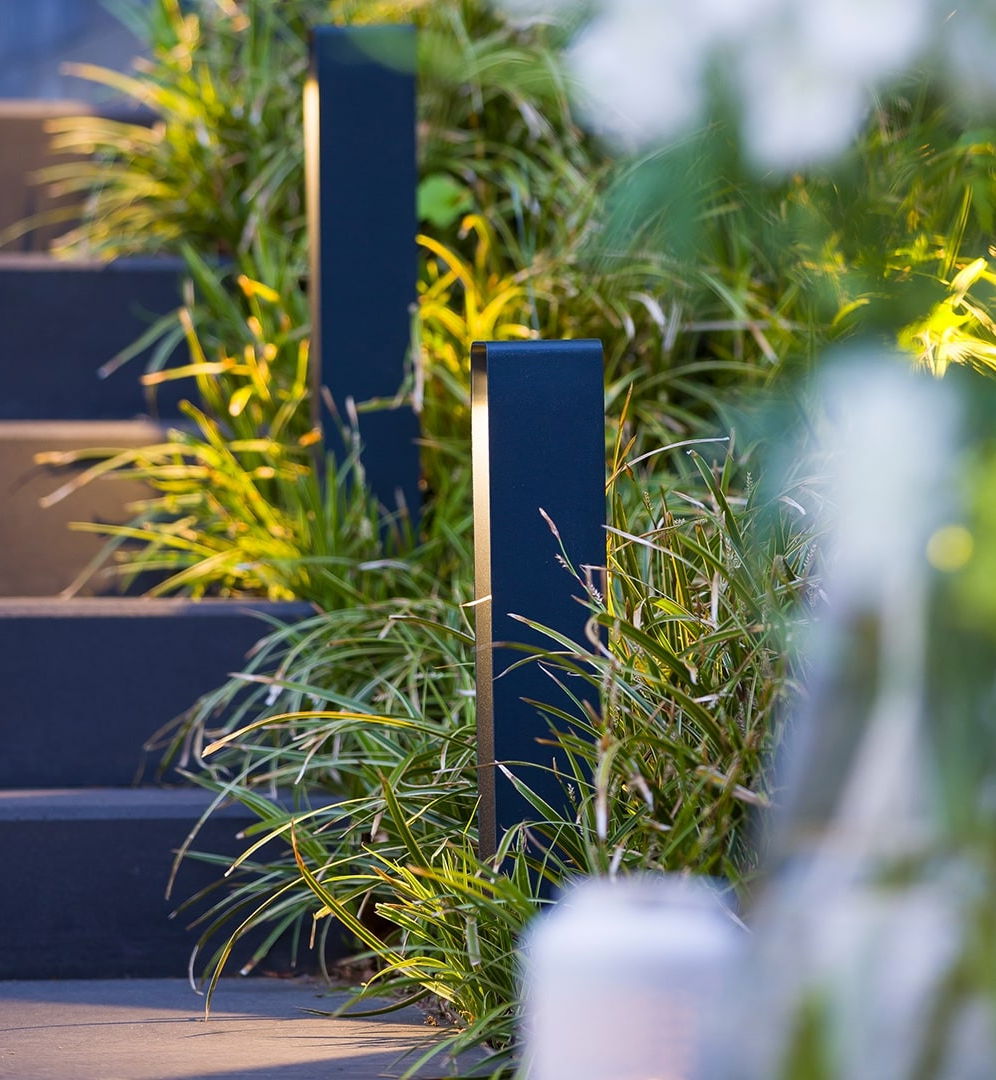 This image depicts a well-designed outdoor lighting fixture integrated into a lush, verdant landscape. The lighting fixture, with its sleek and minimalist black casing, is strategically placed along a set of steps or a raised platform, partially obscured by the surrounding greenery. The vibrant, yellow-green foliage, which appears to be a mix of ornamental grasses and other plant life, frames the lighting fixture and creates a visually striking contrast. The soft, warm glow emitted by the light fixture complements the earthy tones and textures of the plants, resulting in a harmonious and inviting atmosphere. The careful placement of the lighting, combined with the thoughtful landscape design, suggests an intentional approach to blending the built and natural elements seamlessly. The overall impression is one of elegance, sophistication, and a deep appreciation for the interplay between artificial illumination and the natural environment. This image showcases the power of thoughtful outdoor lighting design to enhance the aesthetic and functional qualities of a landscaped space. The integration of the lighting fixture within the verdant setting creates a sense of ambiance and atmosphere, inviting the viewer to imagine the experience of inhabiting this beautifully curated outdoor space.