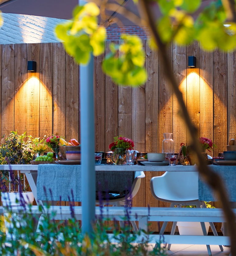 This image depicts a beautifully designed and inviting outdoor dining area, seamlessly blending the natural and built elements to create a warm and welcoming atmosphere.  The focal point is the dining table, which is set with an array of colorful tableware, flowers, and other decorative elements, creating a visually appealing and well-curated display. The table is surrounded by white chairs, adding a touch of minimalism and allowing the natural elements to take center stage.  The wooden fence that lines the perimeter of the space provides a rustic and earthy backdrop, complementing the lush greenery that surrounds the area. The strategically placed lighting fixtures, with their warm, ambient glow, illuminate the space and create a cozy, intimate ambiance as the evening approaches.  The integration of the natural foliage, the carefully selected decor, and the thoughtful lighting design all contribute to the overall sense of harmony and balance within this outdoor retreat. The attention to detail and the seamless blending of the indoor and outdoor elements create an inviting and visually captivating dining experience.  This image showcases the potential of a well-designed outdoor living space, where the boundaries between the built and natural environments are dissolved, allowing the user to fully immerse themselves in a refined and visually stunning setting.