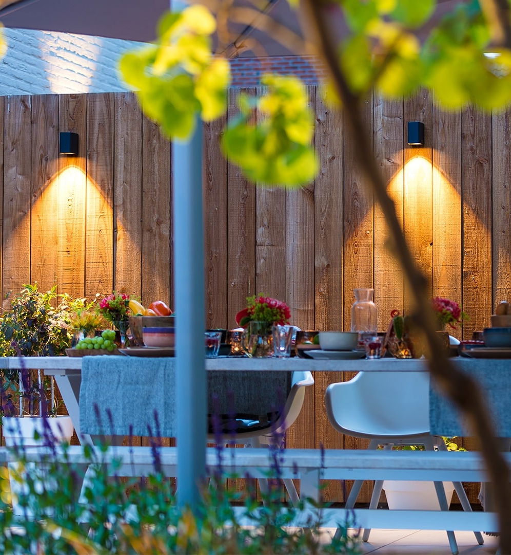 This image depicts a beautifully designed and inviting outdoor dining area, seamlessly blending the natural and built elements to create a warm and welcoming atmosphere. The focal point is the dining table, which is set with an array of colorful tableware, flowers, and other decorative elements, creating a visually appealing and well-curated display. The table is surrounded by white chairs, adding a touch of minimalism and allowing the natural elements to take center stage. The wooden fence that lines the perimeter of the space provides a rustic and earthy backdrop, complementing the lush greenery that surrounds the area. The strategically placed lighting fixtures, with their warm, ambient glow, illuminate the space and create a cozy, intimate ambiance as the evening approaches. The integration of the natural foliage, the carefully selected decor, and the thoughtful lighting design all contribute to the overall sense of harmony and balance within this outdoor retreat. The attention to detail and the seamless blending of the indoor and outdoor elements create an inviting and visually captivating dining experience. This image showcases the potential of a well-designed outdoor living space, where the boundaries between the built and natural environments are dissolved, allowing the user to fully immerse themselves in a refined and visually stunning setting.