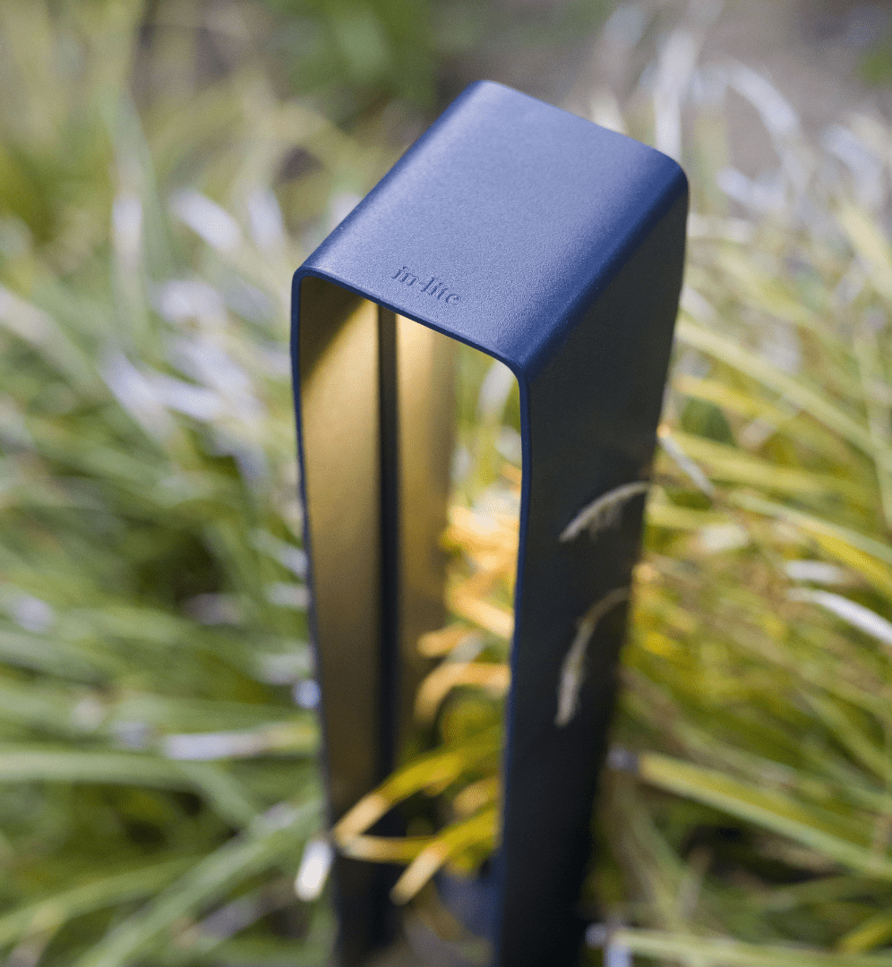 This image depicts a close-up view of a sleek, modern outdoor lighting fixture. The fixture is rectangular in shape, with a matte black or dark blue finish that blends seamlessly with the surrounding natural environment. The fixture is positioned in a lush, grassy area, with blurred greenery and foliage visible in the background, creating a serene and peaceful setting. The lighting fixture appears to be a high-quality, contemporary design, with a minimalist and refined aesthetic. The simple, geometric shape and clean lines of the fixture suggest a focus on form and function, designed to provide illumination while complementing the natural landscape. The name "In Lite" is clearly visible on the side of the fixture, indicating this is a product from a specific manufacturer or brand specializing in outdoor lighting solutions. The overall composition of the image emphasizes the thoughtful integration of the lighting fixture within the natural surroundings, creating a harmonious and visually appealing outdoor environment. This image conveys a sense of modern, minimalist design blended seamlessly with the natural world, highlighting the versatility and visual appeal of well-designed outdoor lighting fixtures that can enhance and complement a range of outdoor settings.