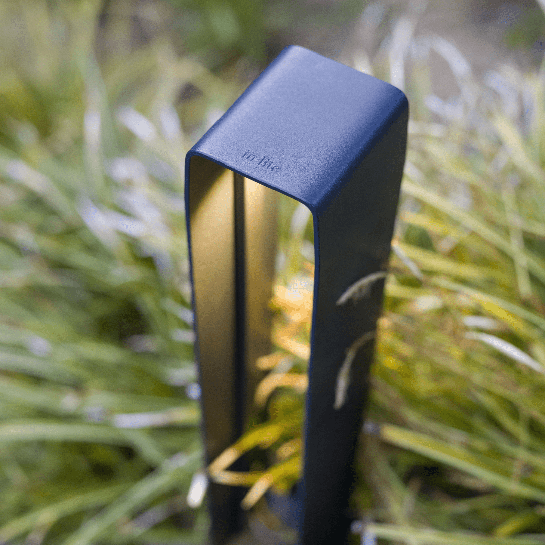 This image depicts a close-up view of a sleek, modern outdoor lighting fixture. The fixture is rectangular in shape, with a matte black or dark blue finish that blends seamlessly with the surrounding natural environment. The fixture is positioned in a lush, grassy area, with blurred greenery and foliage visible in the background, creating a serene and peaceful setting. The lighting fixture appears to be a high-quality, contemporary design, with a minimalist and refined aesthetic. The simple, geometric shape and clean lines of the fixture suggest a focus on form and function, designed to provide illumination while complementing the natural landscape. The name "In Lite" is clearly visible on the side of the fixture, indicating this is a product from a specific manufacturer or brand specializing in outdoor lighting solutions. The overall composition of the image emphasizes the thoughtful integration of the lighting fixture within the natural surroundings, creating a harmonious and visually appealing outdoor environment. This image conveys a sense of modern, minimalist design blended seamlessly with the natural world, highlighting the versatility and visual appeal of well-designed outdoor lighting fixtures that can enhance and complement a range of outdoor settings.