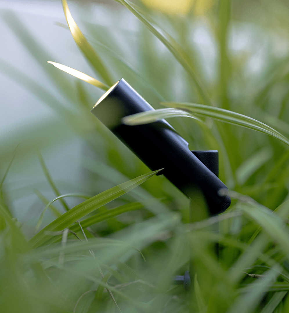 This image depicts a black, cylindrical outdoor lighting fixture discreetly integrated within a lush, verdant natural setting. The fixture is partially obscured by the surrounding foliage, blending seamlessly with the organic elements. The interplay of the artificial lighting component and the vibrant greenery creates a sense of harmony and balance, showcasing how in-lite's lighting solutions can be thoughtfully incorporated into outdoor environments. The image suggests a focus on sustainability and considerate design, where the lighting enhances the beauty of nature without overpowering or disrupting the natural surroundings.