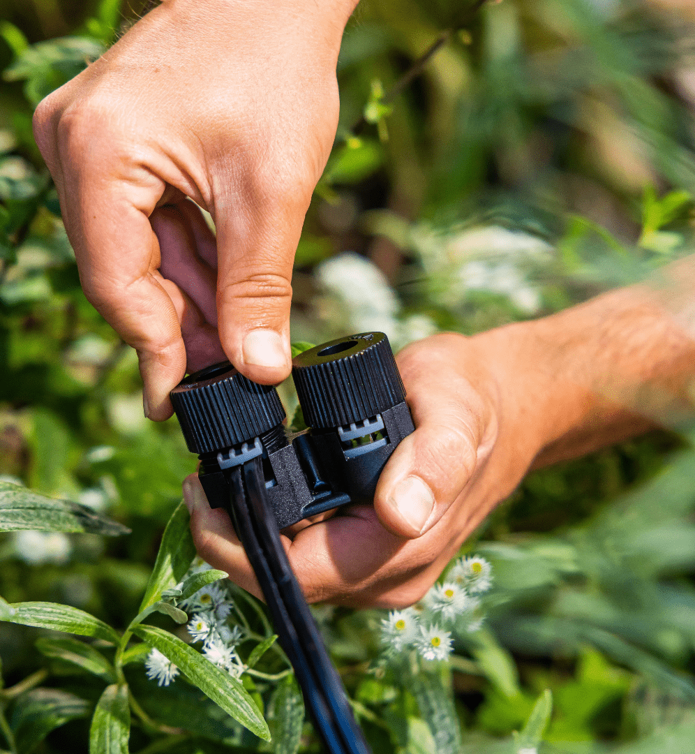 De afbeelding laat iemands handen zien die een zwart, elektrisch gereedschap vastpakken dat lijkt te worden gebruikt voor tuinonderhoud of gewasverzorging. De achtergrond bestaat uit groen gebladerte en bloeiende, witte bloemen, wat suggereert dat het gereedschap wordt gebruikt in een natuurlijke, buitenomgeving. De focus en compositie van de afbeelding leggen de nadruk op de interactie tussen de menselijke handen en het technologische hulpmiddel, waarbij de natuur op de achtergrond een rustgevend contrast vormt.