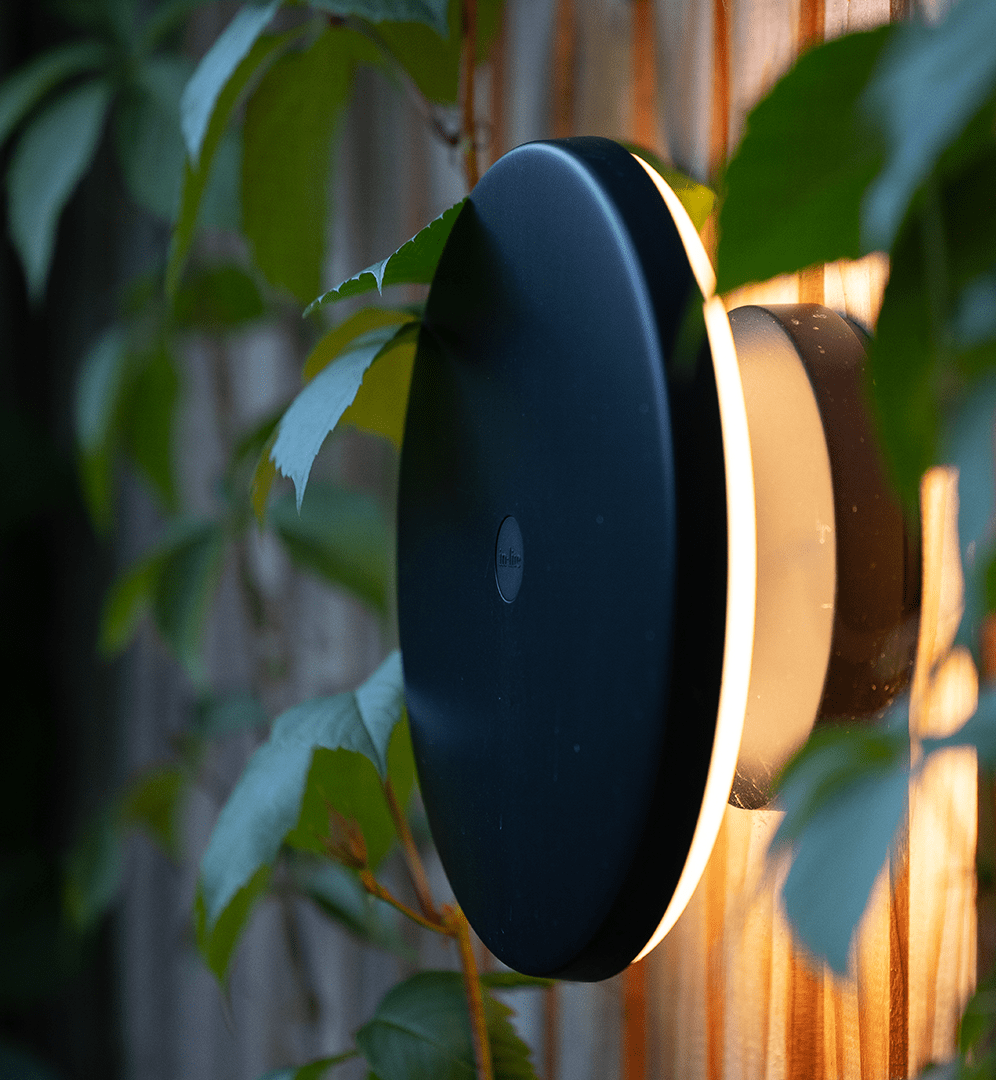 This image depicts a sleek, modern outdoor lighting fixture that is integrated into a lush, natural setting. The fixture has a rounded, disc-shaped design with a glossy black exterior and a warm, golden-toned lighting element at its center. The lighting fixture is mounted on a wooden surface or structure, blending seamlessly with the surrounding greenery. The vibrant, verdant foliage and leaves in the background create a striking contrast with the dark, minimalist design of the light. The overall composition highlights the harmonious integration of the designed, technological element with the organic, living components of the garden environment. The subtle, ambient glow emanating from the fixture adds a touch of warmth and elegance to the natural setting, creating a visually striking and cohesive aesthetic. This image conveys a sense of thoughtful, modern design that elevates the functional purpose of outdoor lighting into a visually captivating and environmentally-conscious artistic statement, where the man-made and the natural coexist in a harmonious and aesthetically pleasing manner.