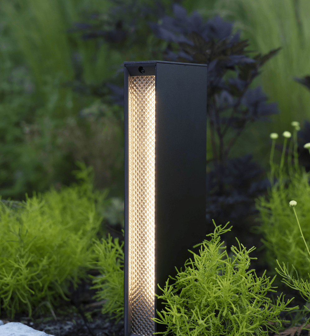 This image depicts a modern, minimalist outdoor lighting fixture positioned amidst a lush, naturalistic garden setting. The lighting fixture is a tall, rectangular shape with a matte black exterior and a central column of warm, diffused lighting. The fixture appears to be a high-quality, architectural lighting solution, designed to blend seamlessly with the surrounding natural environment. The textured, perforated surface of the central column creates a visually interesting and unique lighting effect, casting a soft, ambient glow that highlights the vibrant greens and textures of the nearby foliage. The fixture is situated on a paved path or walkway, with the lush, verdant plants and shrubs surrounding it creating a sense of depth and immersion within the outdoor space. The overall composition emphasizes the harmonious integration of the designed lighting element and the natural landscape, creating a cohesive and visually striking aesthetic. This image conveys a sense of thoughtful, intentional design that enhances and complements the inherent beauty of the outdoor setting, elevating the functionality of the lighting fixture into a design statement that seamlessly blends the man-made and the natural.