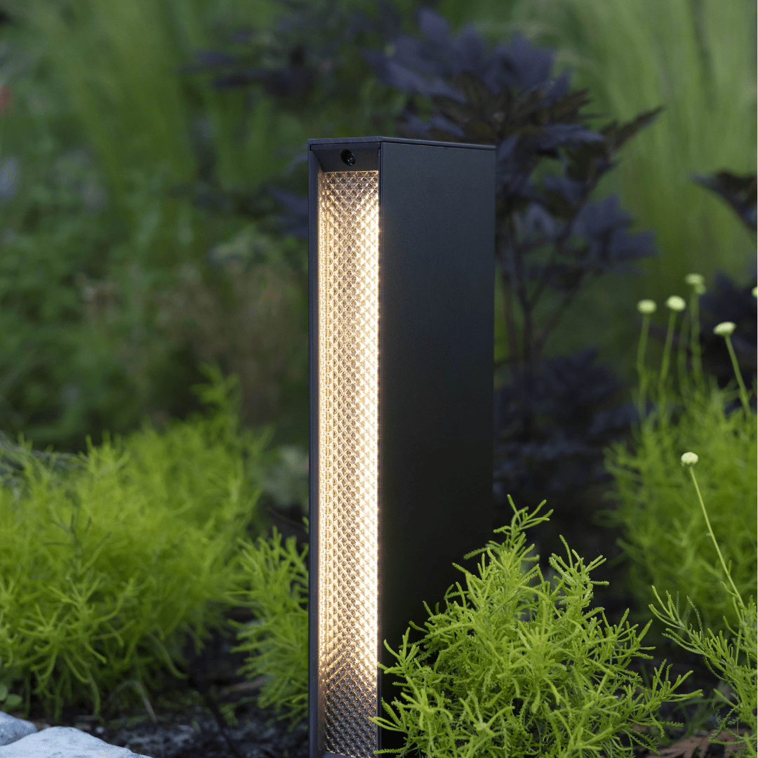 This image depicts a modern, minimalist outdoor lighting fixture positioned amidst a lush, naturalistic garden setting. The lighting fixture is a tall, rectangular shape with a matte black exterior and a central column of warm, diffused lighting. The fixture appears to be a high-quality, architectural lighting solution, designed to blend seamlessly with the surrounding natural environment. The textured, perforated surface of the central column creates a visually interesting and unique lighting effect, casting a soft, ambient glow that highlights the vibrant greens and textures of the nearby foliage. The fixture is situated on a paved path or walkway, with the lush, verdant plants and shrubs surrounding it creating a sense of depth and immersion within the outdoor space. The overall composition emphasizes the harmonious integration of the designed lighting element and the natural landscape, creating a cohesive and visually striking aesthetic. This image conveys a sense of thoughtful, intentional design that enhances and complements the inherent beauty of the outdoor setting, elevating the functionality of the lighting fixture into a design statement that seamlessly blends the man-made and the natural.