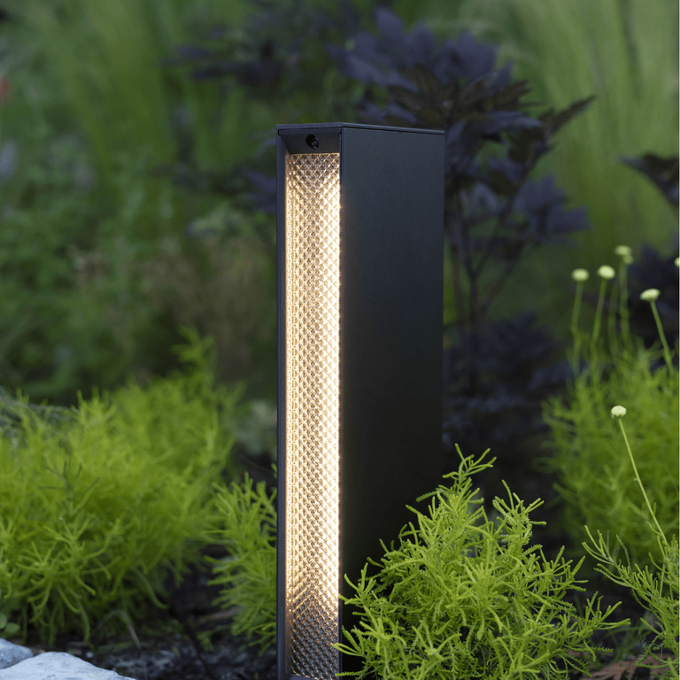 This image depicts a modern, minimalist outdoor lighting fixture positioned amidst a lush, naturalistic garden setting. The lighting fixture is a tall, rectangular shape with a matte black exterior and a central column of warm, diffused lighting. The fixture appears to be a high-quality, architectural lighting solution, designed to blend seamlessly with the surrounding natural environment. The textured, perforated surface of the central column creates a visually interesting and unique lighting effect, casting a soft, ambient glow that highlights the vibrant greens and textures of the nearby foliage. The fixture is situated on a paved path or walkway, with the lush, verdant plants and shrubs surrounding it creating a sense of depth and immersion within the outdoor space. The overall composition emphasizes the harmonious integration of the designed lighting element and the natural landscape, creating a cohesive and visually striking aesthetic. This image conveys a sense of thoughtful, intentional design that enhances and complements the inherent beauty of the outdoor setting, elevating the functionality of the lighting fixture into a design statement that seamlessly blends the man-made and the natural.