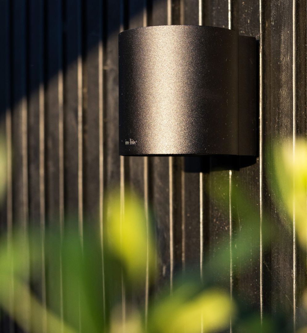 This image depicts a dark, moody outdoor scene with a focus on a weathered wooden fence and a single outdoor lighting fixture. The lighting fixture, which appears to be a wall-mounted sconce, is the primary focal point, with its sleek, matte black design standing out against the weathered wooden slats of the fence. The lighting fixture is casting a soft, warm glow, which creates a sense of depth and atmosphere in the surrounding area. The blurred, out-of-focus greenery in the foreground suggests a lush, overgrown garden or natural environment surrounding the fence, adding to the overall sense of enclosure and intimacy in the scene. The contrasting textures and tones create a moody, contemplative mood, inviting the viewer to pause and appreciate the interplay of light, shadow, and natural elements in this carefully composed outdoor setting.