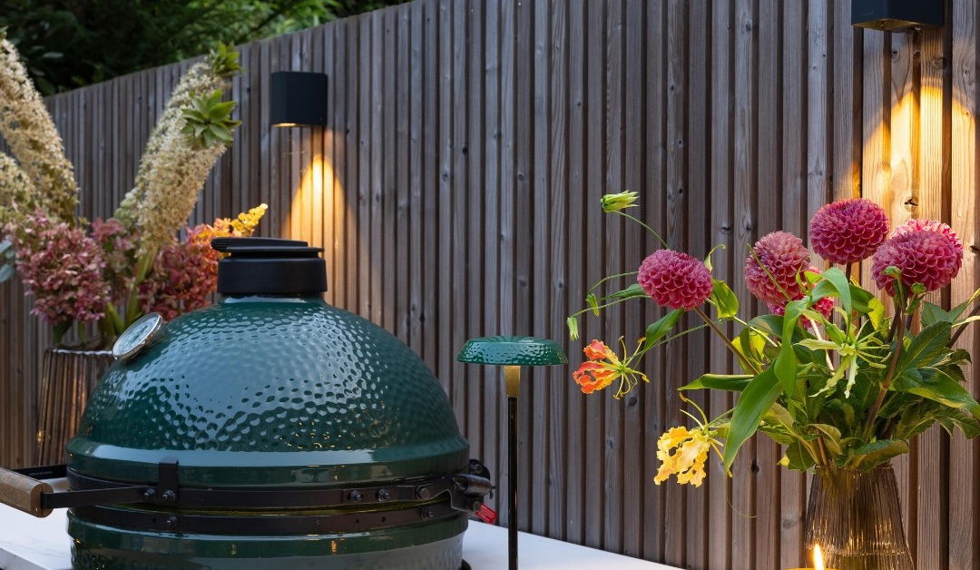 This image depicts a cozy and inviting outdoor cooking setup in a lush, garden-like environment. The focal point is a large, green egg-shaped charcoal grill or smoker, which is surrounded by a wooden fence and framed by various ornamental plants and flowers. The grill is positioned on a white, minimalist counter or table, and there are small tealight candles or lanterns providing soft, warm lighting. The overall impression is one of a tranquil and well-designed outdoor living space, ideal for entertaining or relaxing while preparing delicious meals.