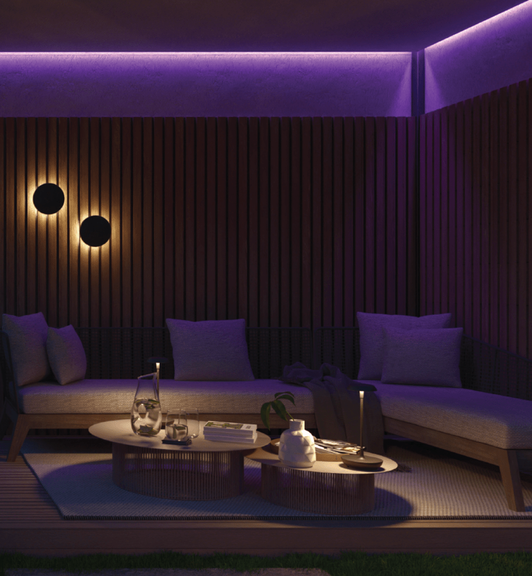 The image depicts a cozy and inviting outdoor living space at night. The focal point is a covered seating area with comfortable-looking purple sofas and chairs, creating a warm and intimate atmosphere. This seating area is illuminated by subtle, warm lighting, including overhead fixtures and wall-mounted lamps, which cast a soothing glow over the space. The surrounding wooden walls and the presence of a lush, mature tree with vibrant foliage add a natural and serene quality to the scene. A coffee table with various decorative elements is also visible, suggesting this area is designed for relaxation and socializing. The overall impression is one of a beautifully designed and thoughtfully curated outdoor retreat, perfectly suited for enjoying the tranquility of the evening.