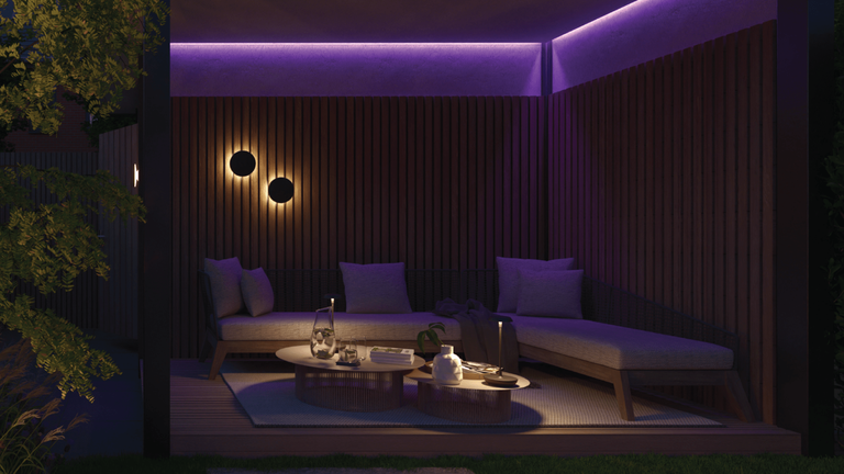 The image depicts a cozy and inviting outdoor living space at night. The focal point is a covered seating area with comfortable-looking purple sofas and chairs, creating a warm and intimate atmosphere. This seating area is illuminated by subtle, warm lighting, including overhead fixtures and wall-mounted lamps, which cast a soothing glow over the space. The surrounding wooden walls and the presence of a lush, mature tree with vibrant foliage add a natural and serene quality to the scene. A coffee table with various decorative elements is also visible, suggesting this area is designed for relaxation and socializing. The overall impression is one of a beautifully designed and thoughtfully curated outdoor retreat, perfectly suited for enjoying the tranquility of the evening.