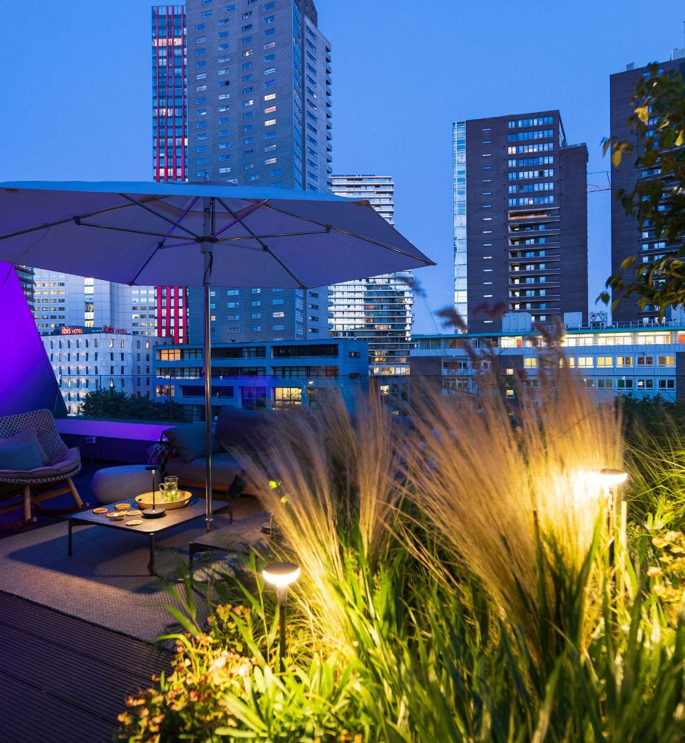 This image depicts a cozy and luxurious outdoor living space in an urban setting. The focal point is a covered seating area with comfortable-looking furniture, including a plush sofa and chairs, situated on a wooden deck. The seating area is illuminated by a large, purple-hued patio umbrella, which creates a warm and inviting ambiance.  The surrounding area is lush with various plants and greenery, including tall, vibrant grasses and other foliage that add to the serene, natural atmosphere. In the background, the city skyline is visible, with a mix of modern high-rise buildings and lighting that creates a striking contrast between the natural and urban elements.  The overall impression is one of a beautifully designed and carefully curated outdoor oasis, where the residents can enjoy the comforts of home while immersed in nature and overlooking the dynamic city landscape. The attention to detail, from the thoughtful lighting to the carefully selected plants, creates a visually captivating and inviting space for relaxation and entertainment.